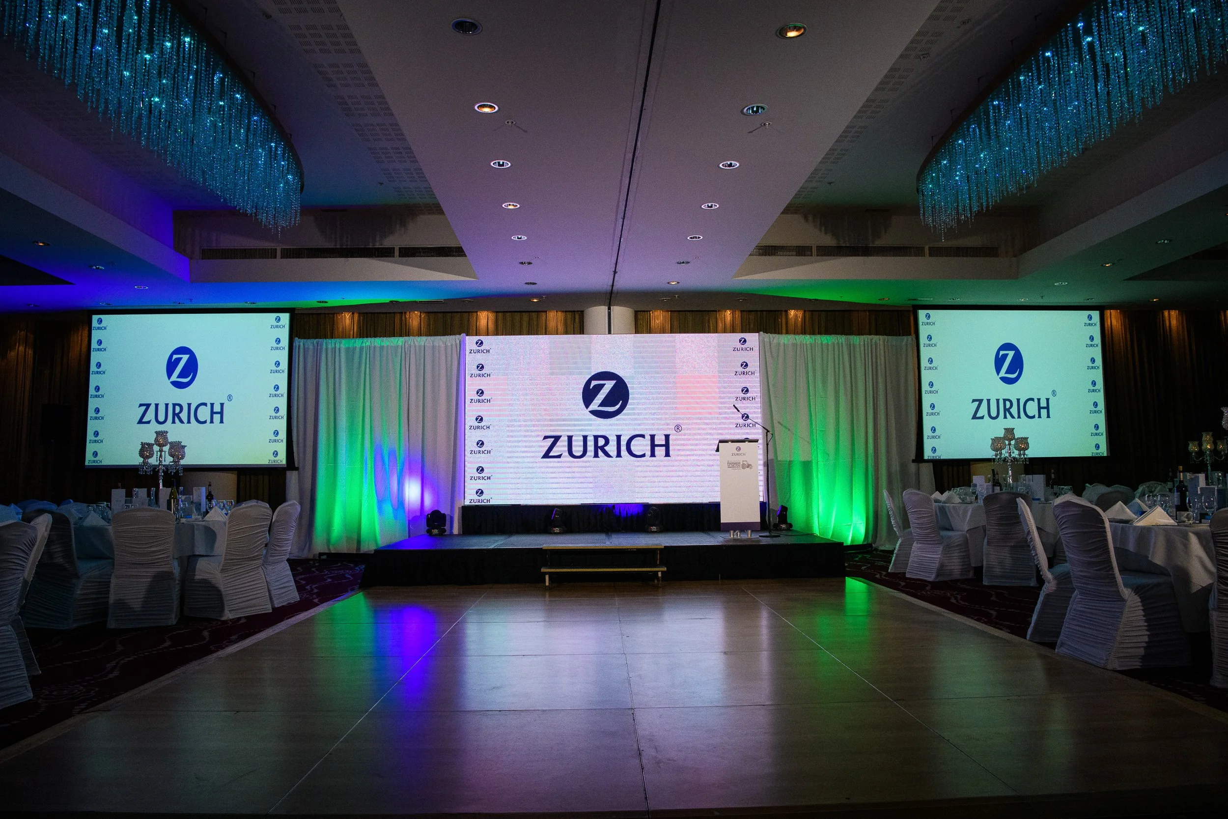 Corporate event hall with stage, LED screens displaying branding, and banquet tables arranged beneath chandeliers.
