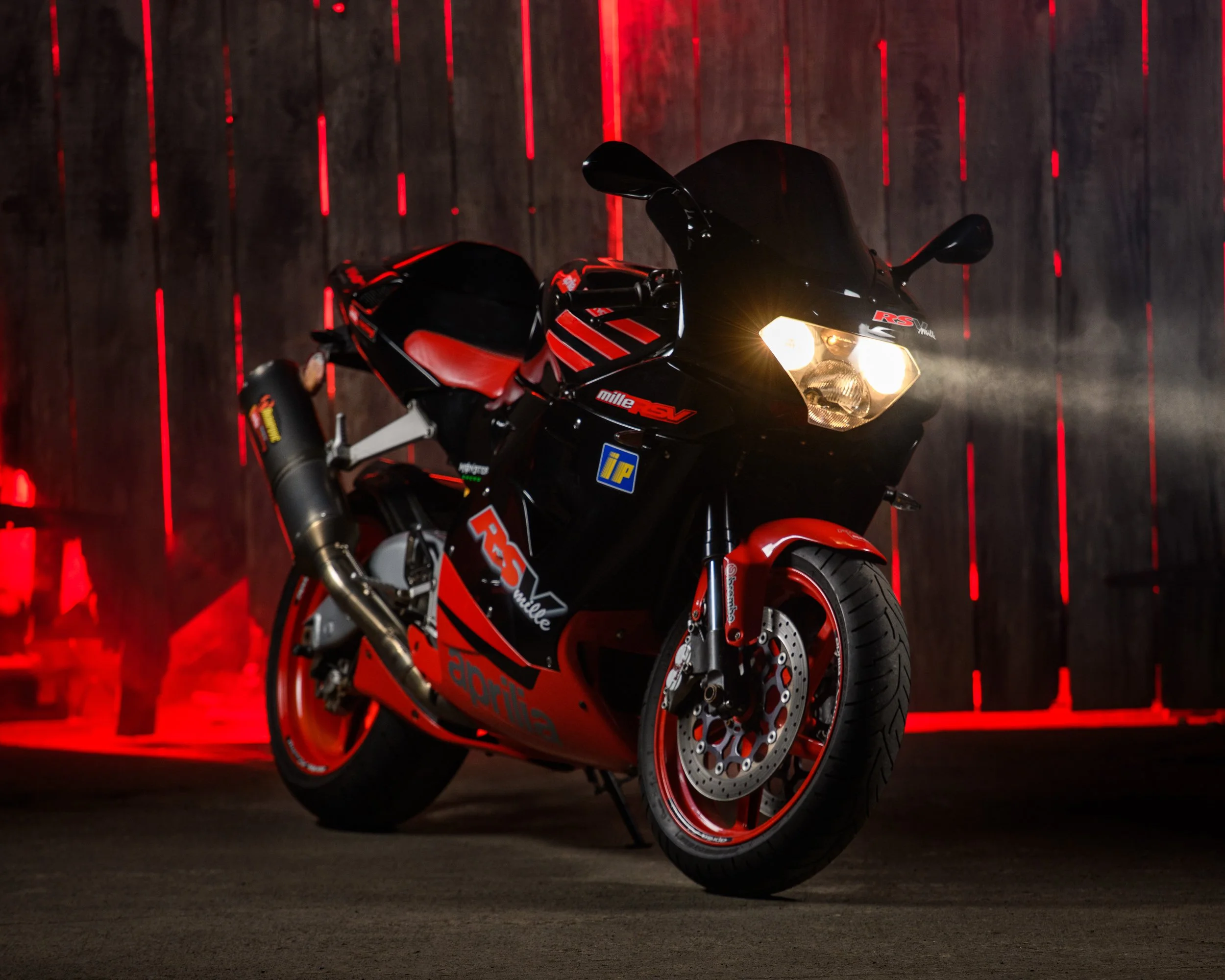 Black and red Aprilia sport motorcycle photographed indoors against a dark wooden backdrop with red accent lighting.
