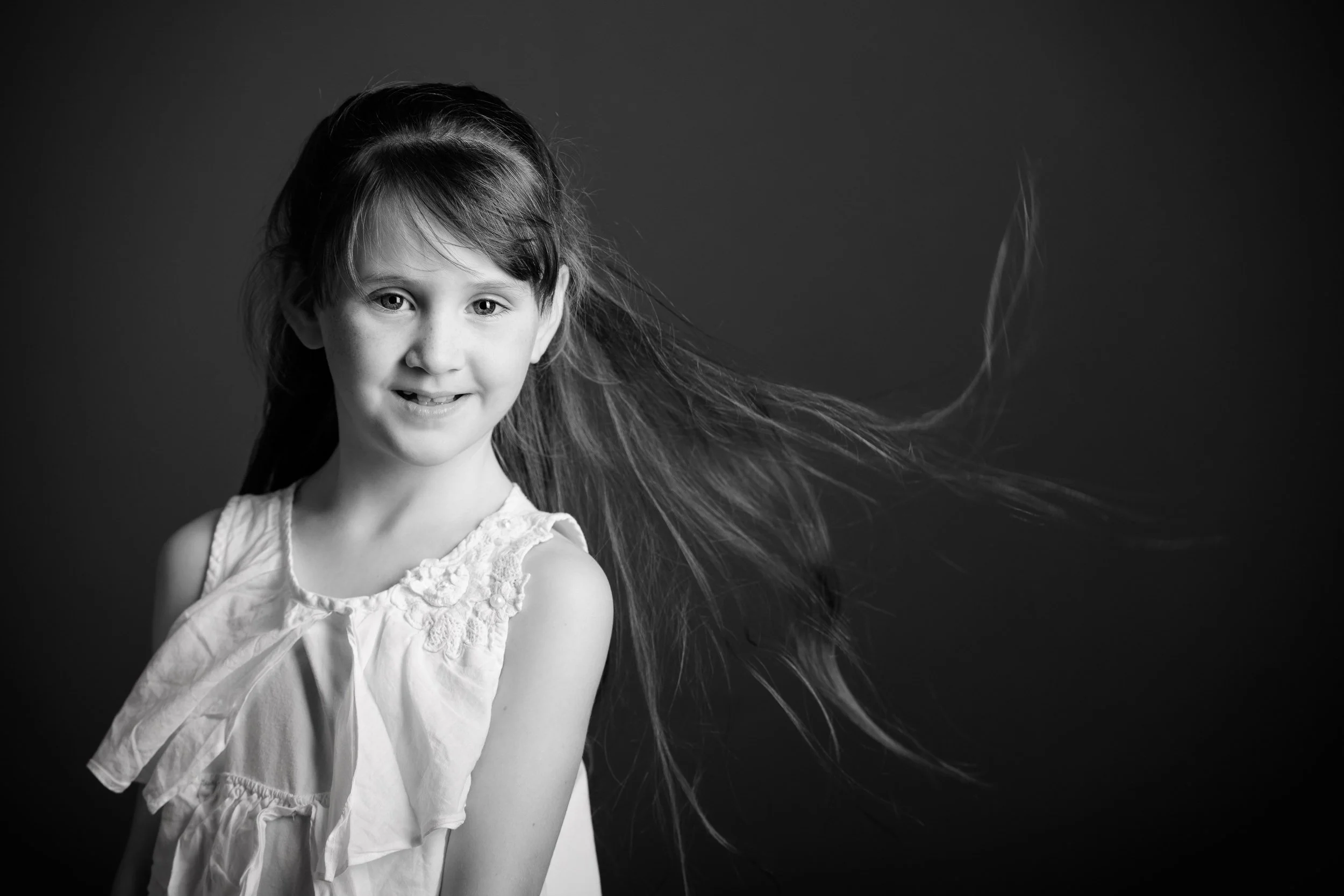 Black and white photo of a young girl with long hair, smiling at the camera, wearing a sleeveless top with lace details, and her hair flowing to the side.