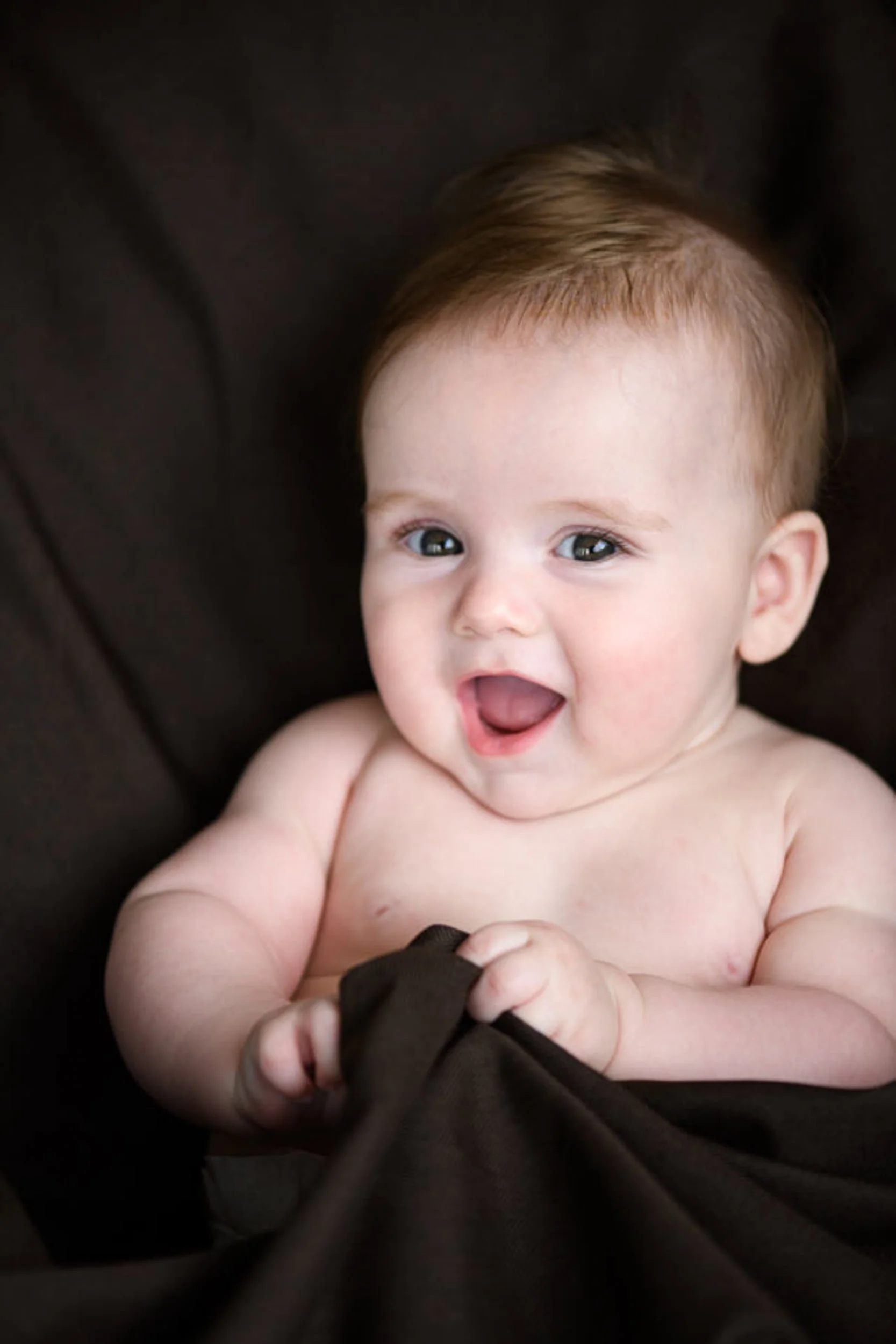 A smiling baby with light skin and brown hair, lying on a dark surface, holding a piece of dark fabric.