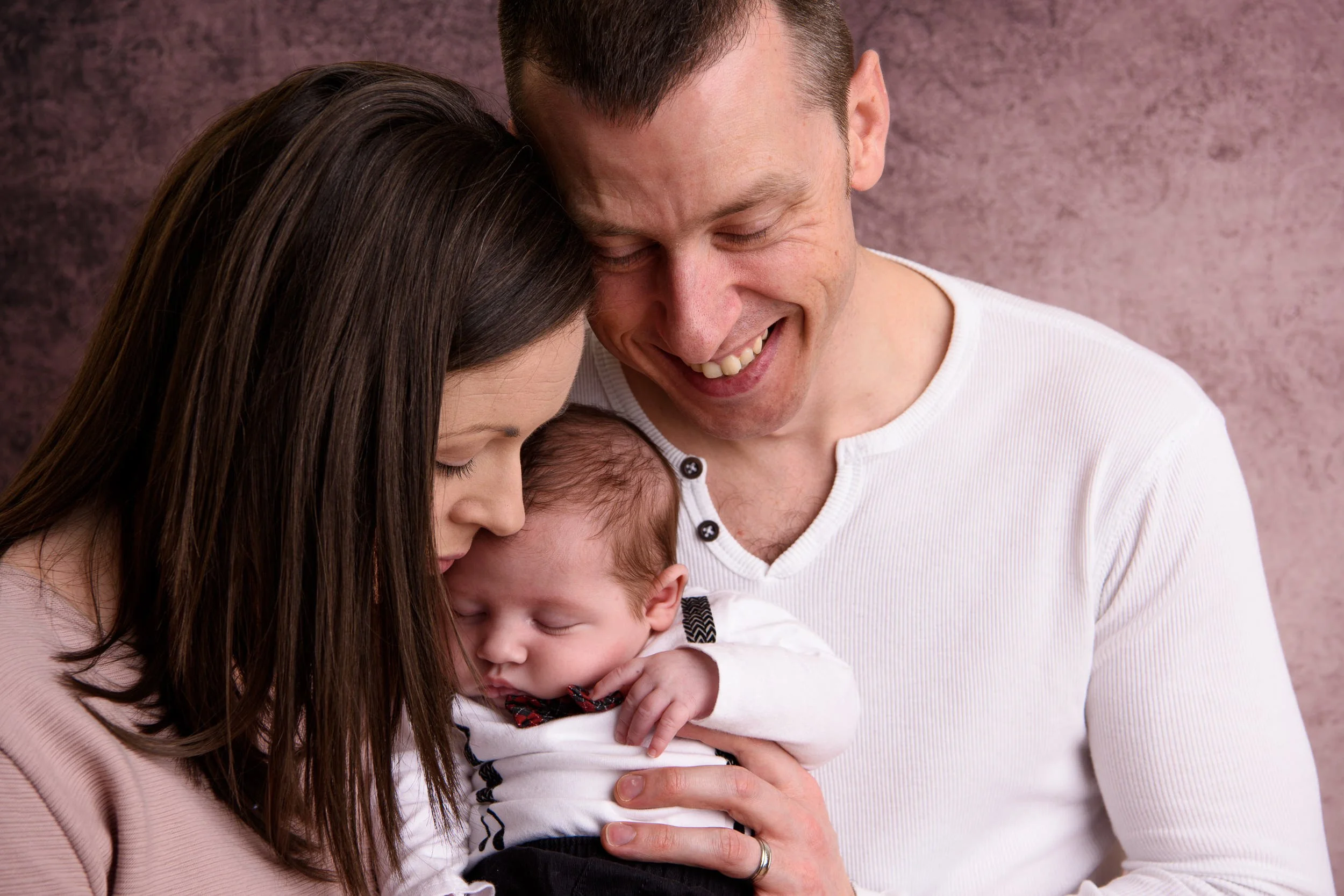 A man holding a sleeping baby while a woman leans in close, sharing a warm family moment against a soft pink background.
