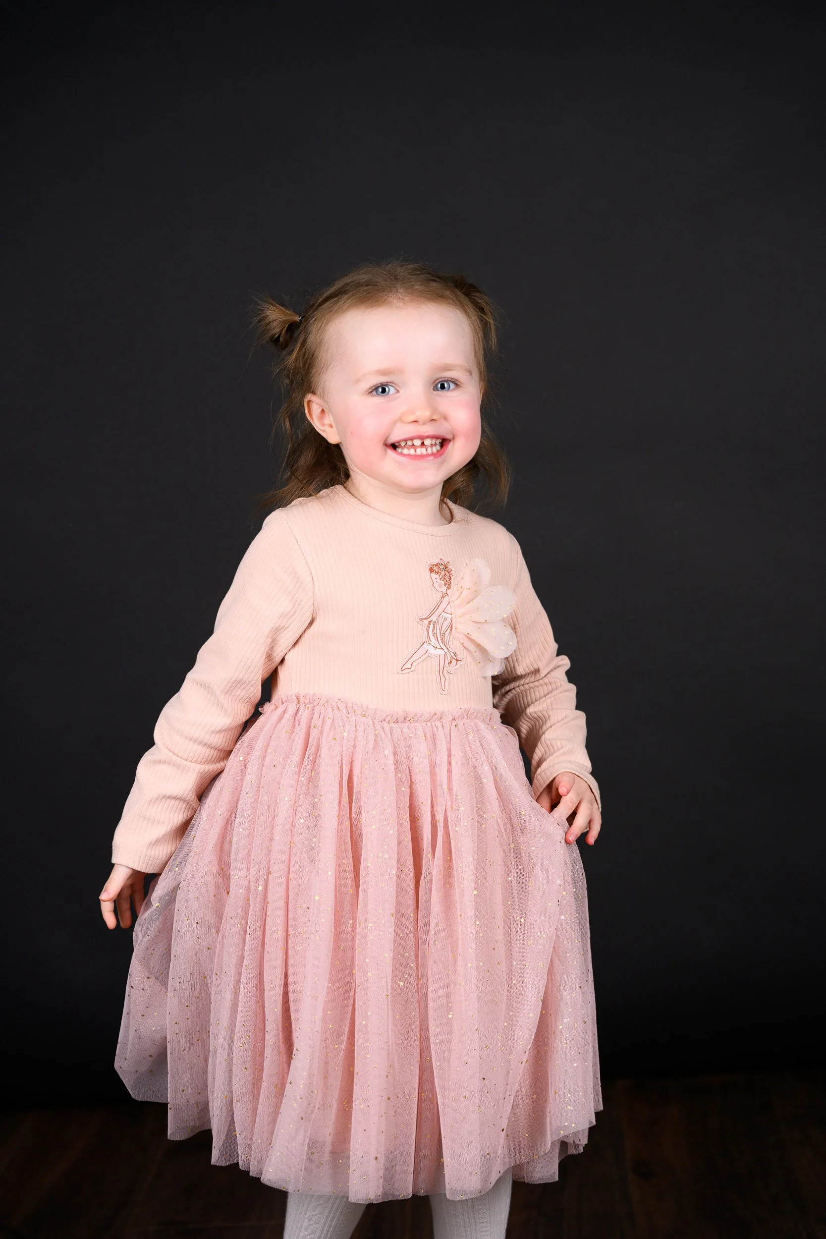 A young girl in a pink dress twirling on a wooden floor against a dark background.
