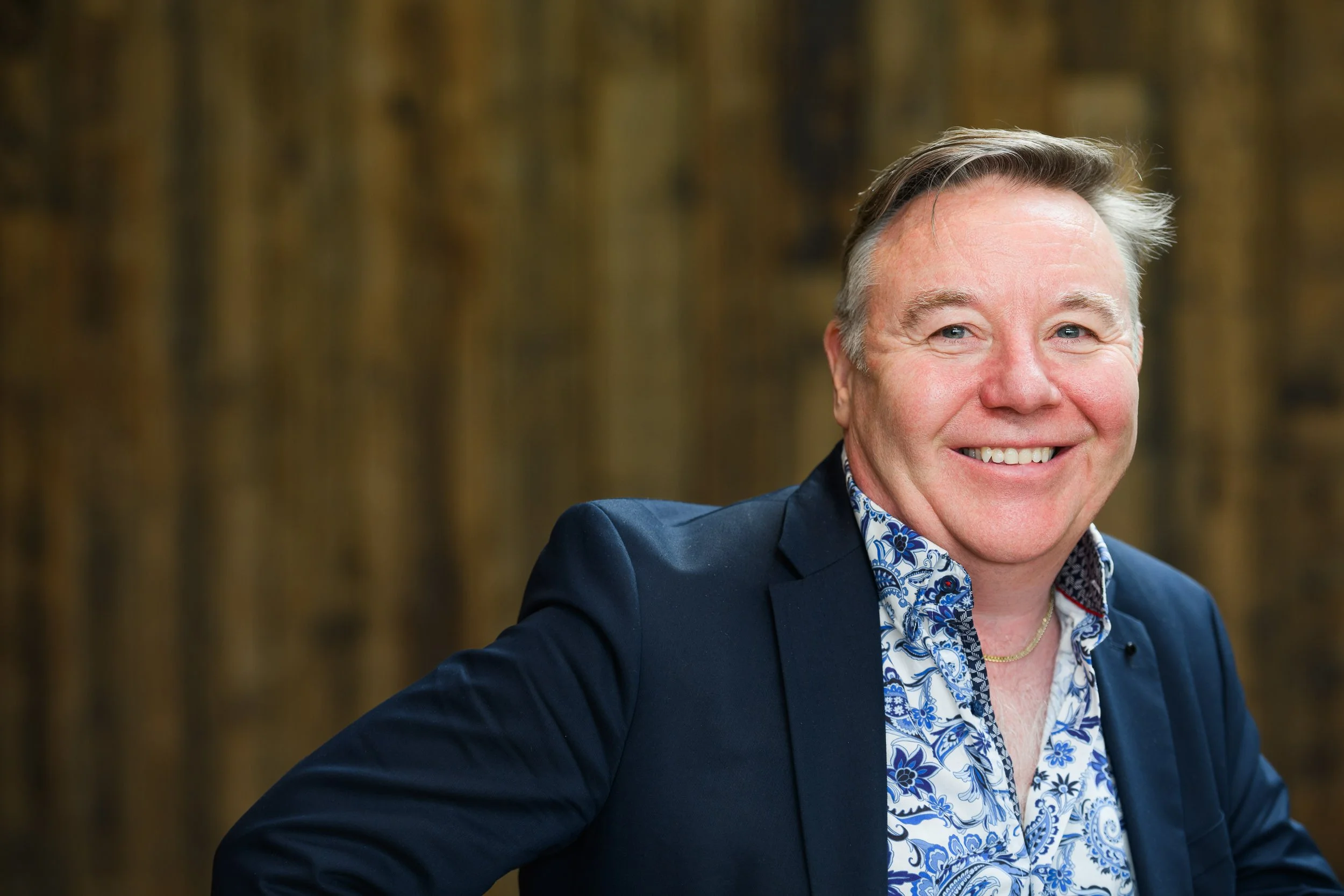 Portrait of a middle-aged man with grey hair wearing a blue blazer, photographed outdoors with a softly blurred natural background.
