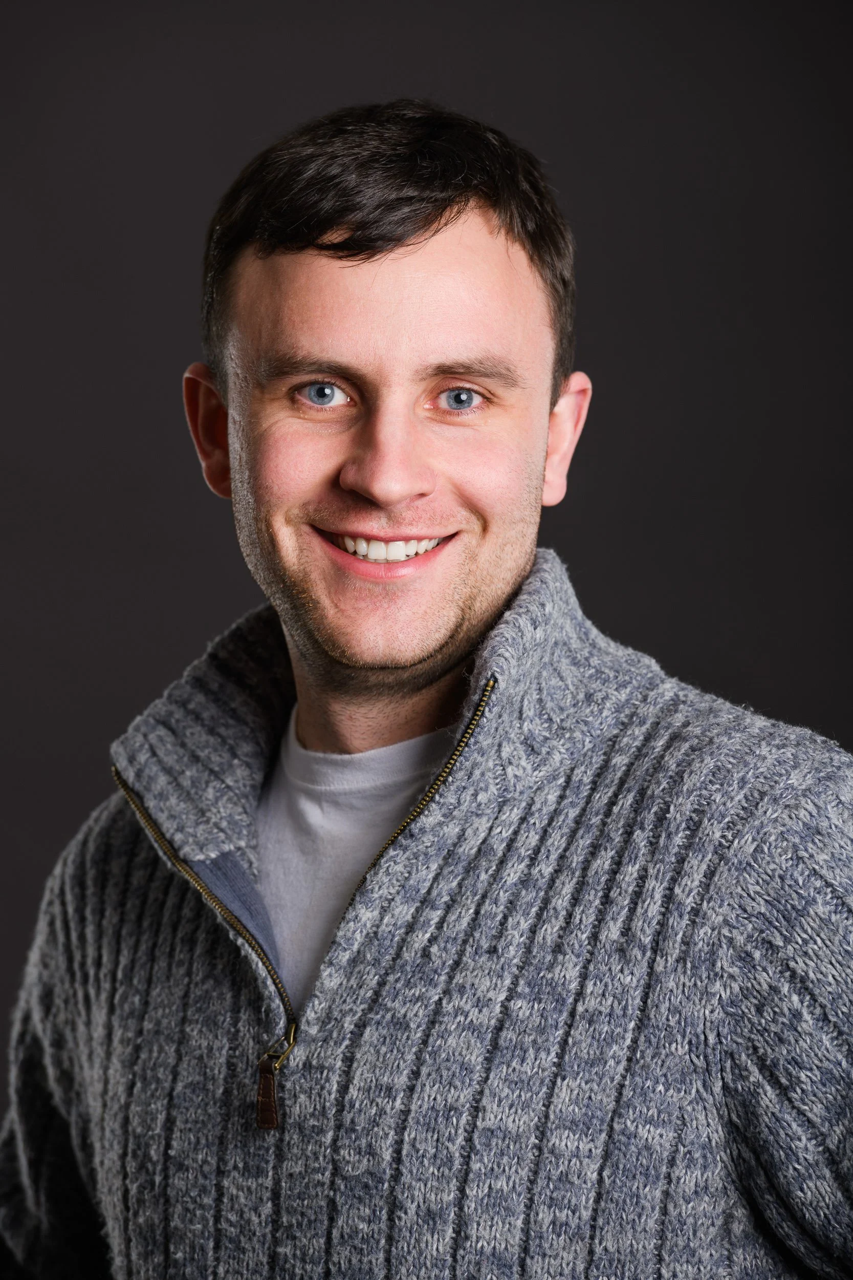 Portrait of a smiling man with short dark hair wearing a grey zip-up sweater over a white t-shirt, against a dark background.
