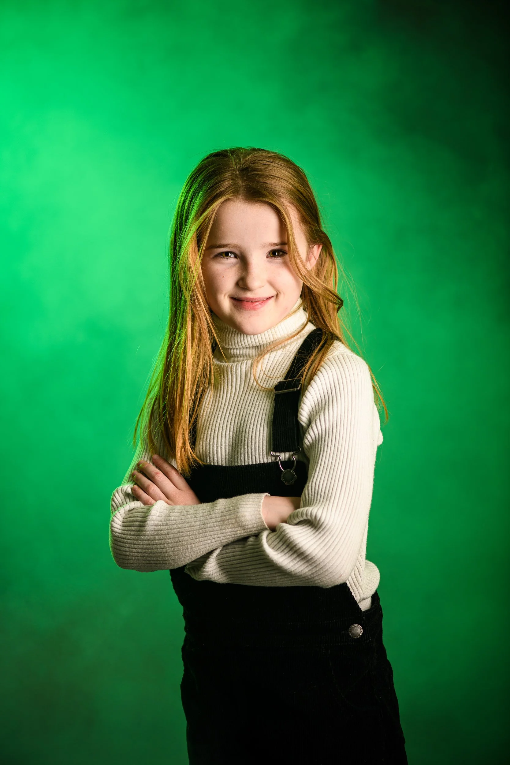 A young girl with long red hair, smiling and crossing her arms, standing against a green background.