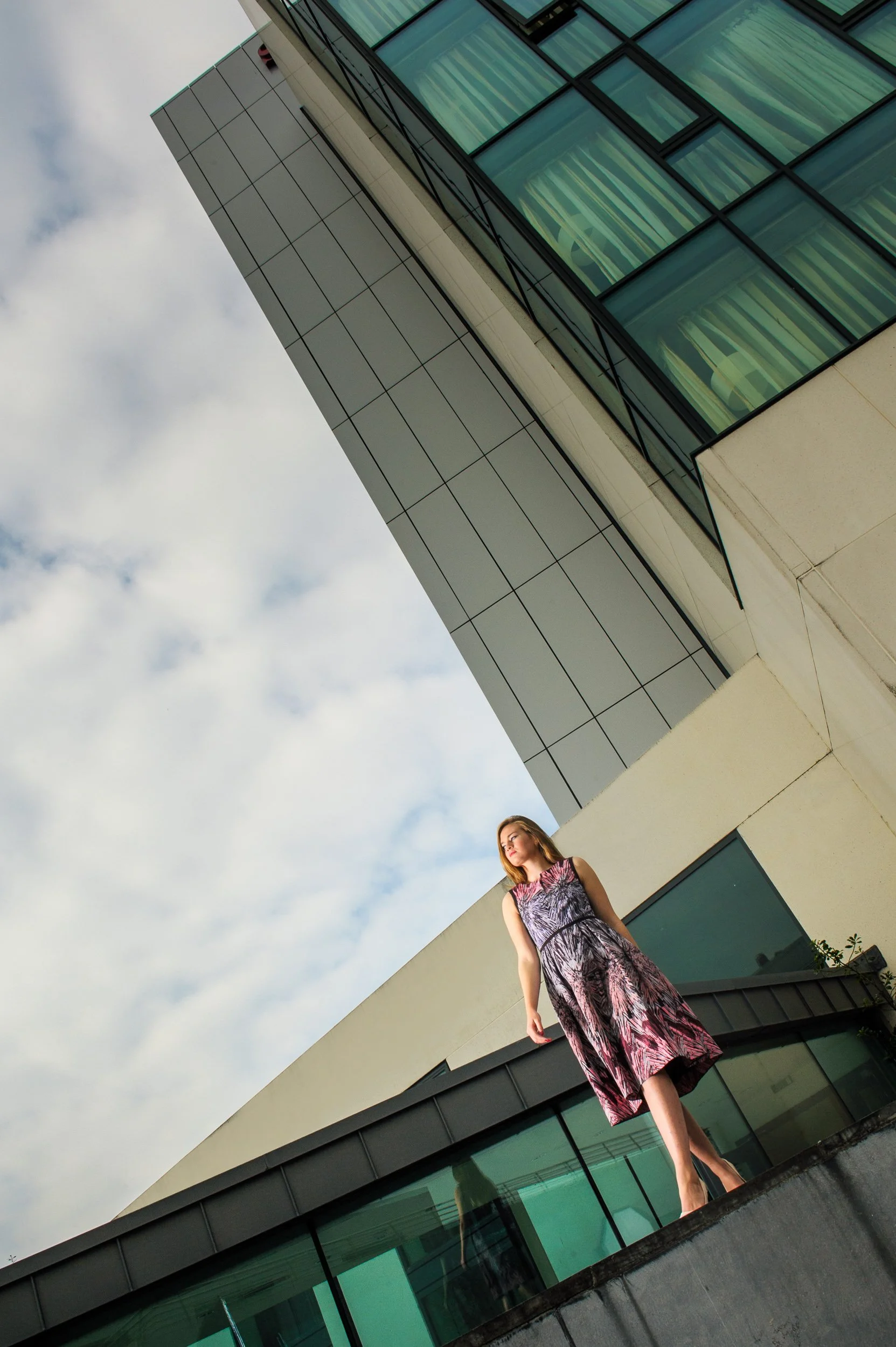 Modern fashion portrait of a woman standing on a ledge outside a glass building under a cloudy sky