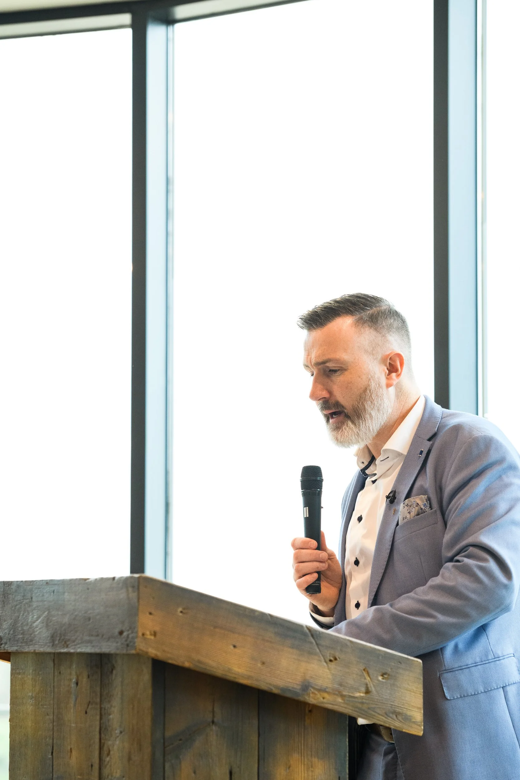 A man in a suit speaking at a podium with a microphone in a bright event space.
