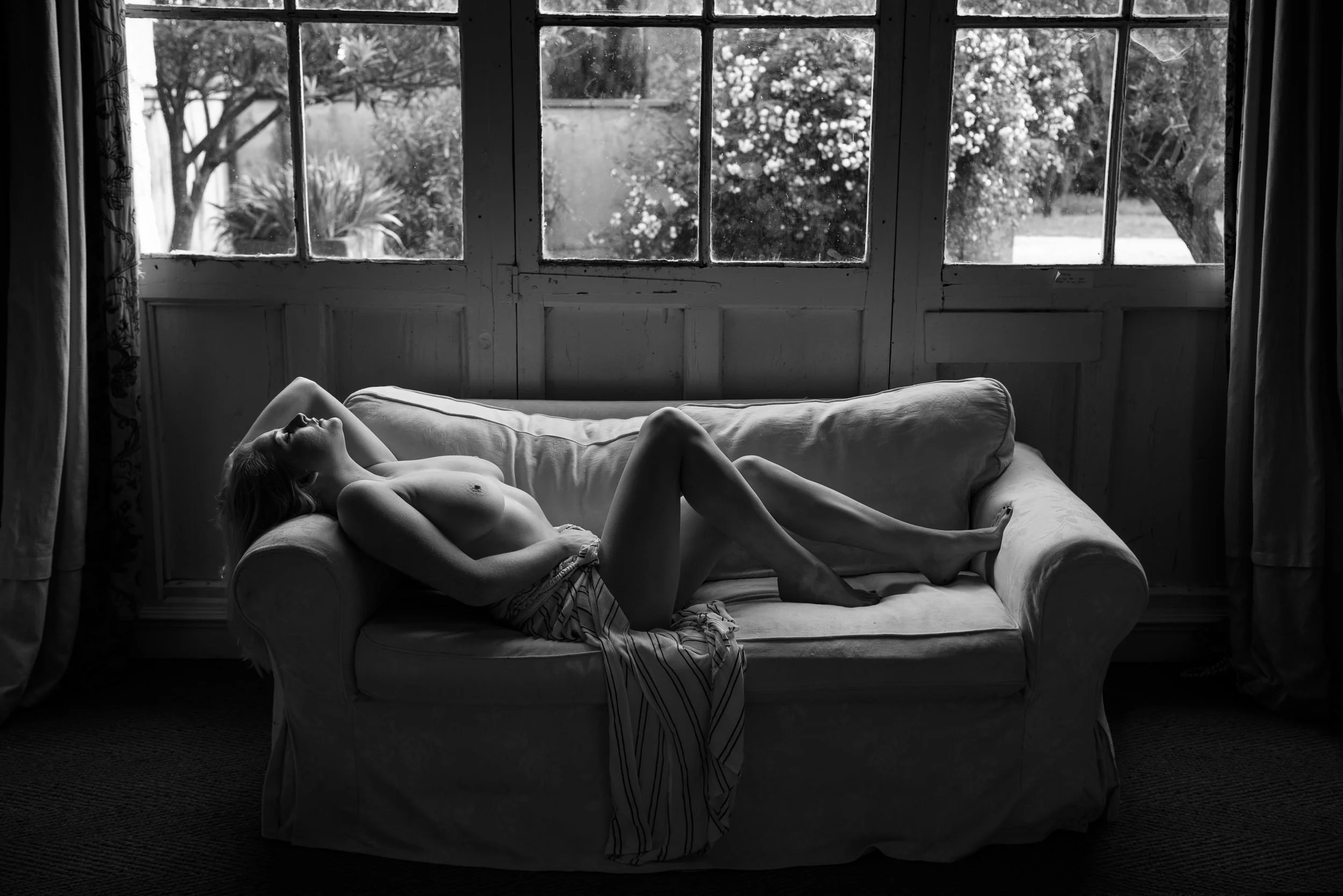 Black and white portrait of a woman reclining on a sofa beside large windows, with soft natural light and greenery beyond.
