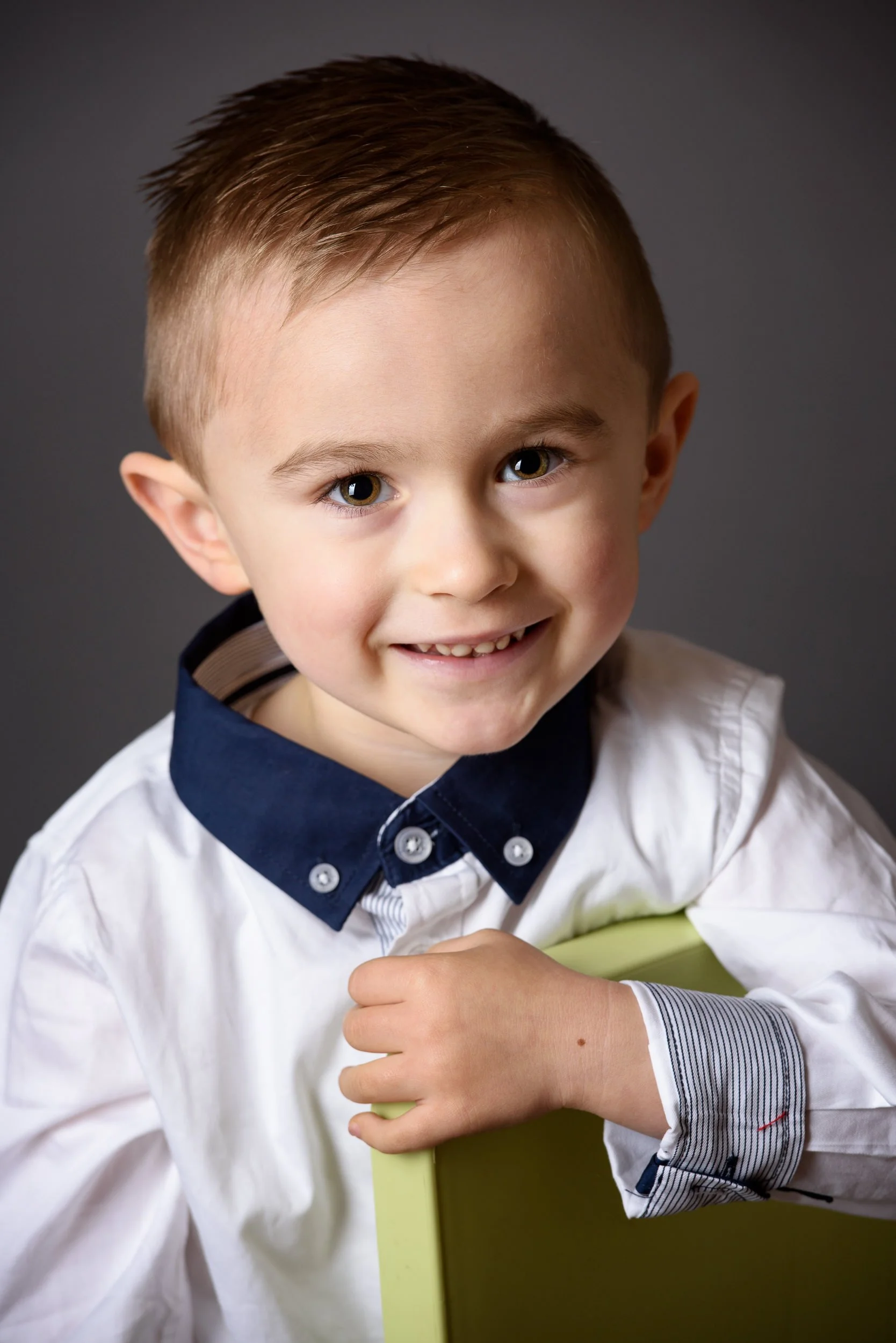 A young boy with brown hair and blue eyes smiling while holding a green box. He is wearing a white shirt with a dark blue collar with white buttons and striped cuffs.