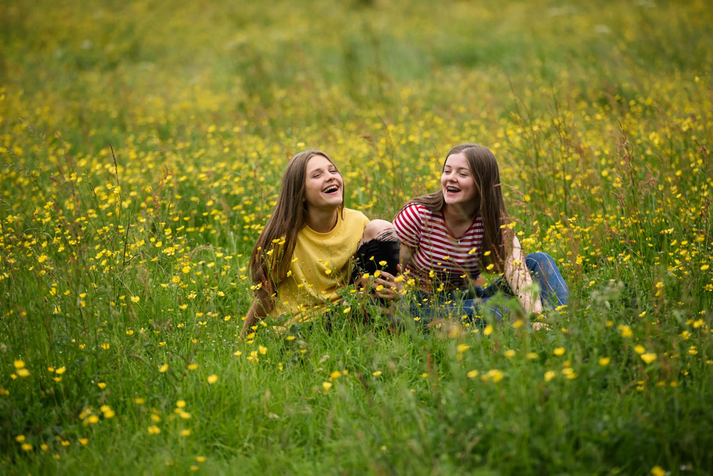 Two young women sitting in a field of yellow flowers, laughing and enjoying each other's company.