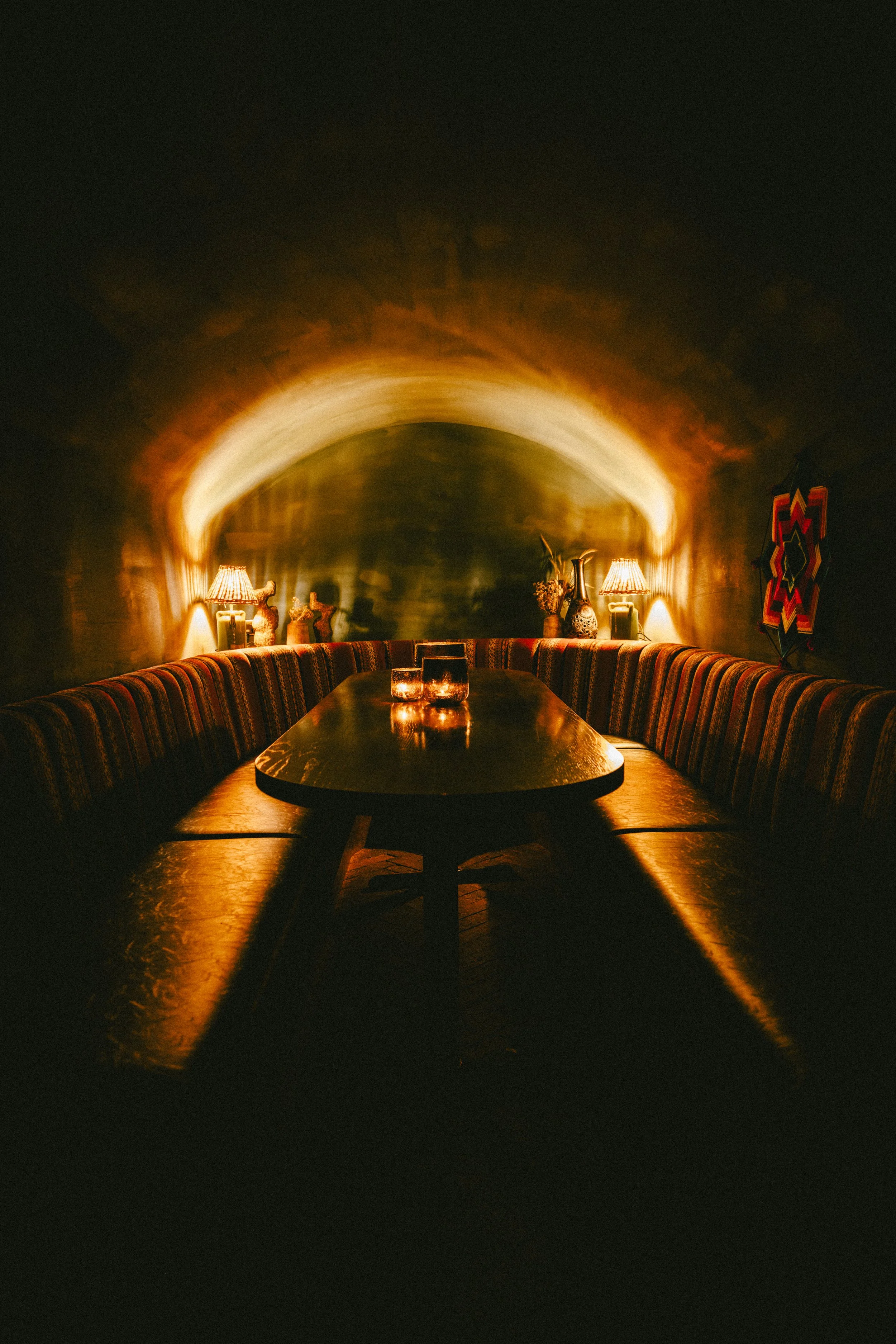 Dimly lit room with a long wooden table, surrounded by a curved upholstered bench. Warm lamps and decorative vases on the side, with artwork on the wall. Focus on ambient lighting and cozy atmosphere.