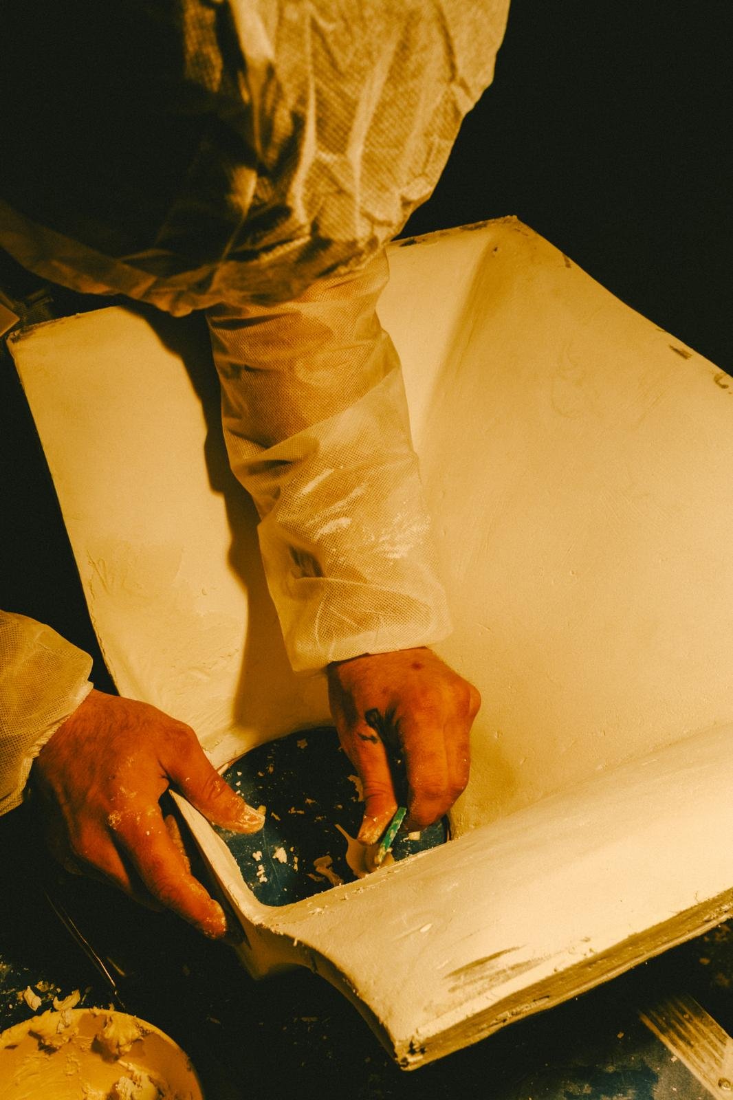 A person wearing a beige jacket is scooping ice cream from a container into a bowl with an spoon. The scene is dimly lit.