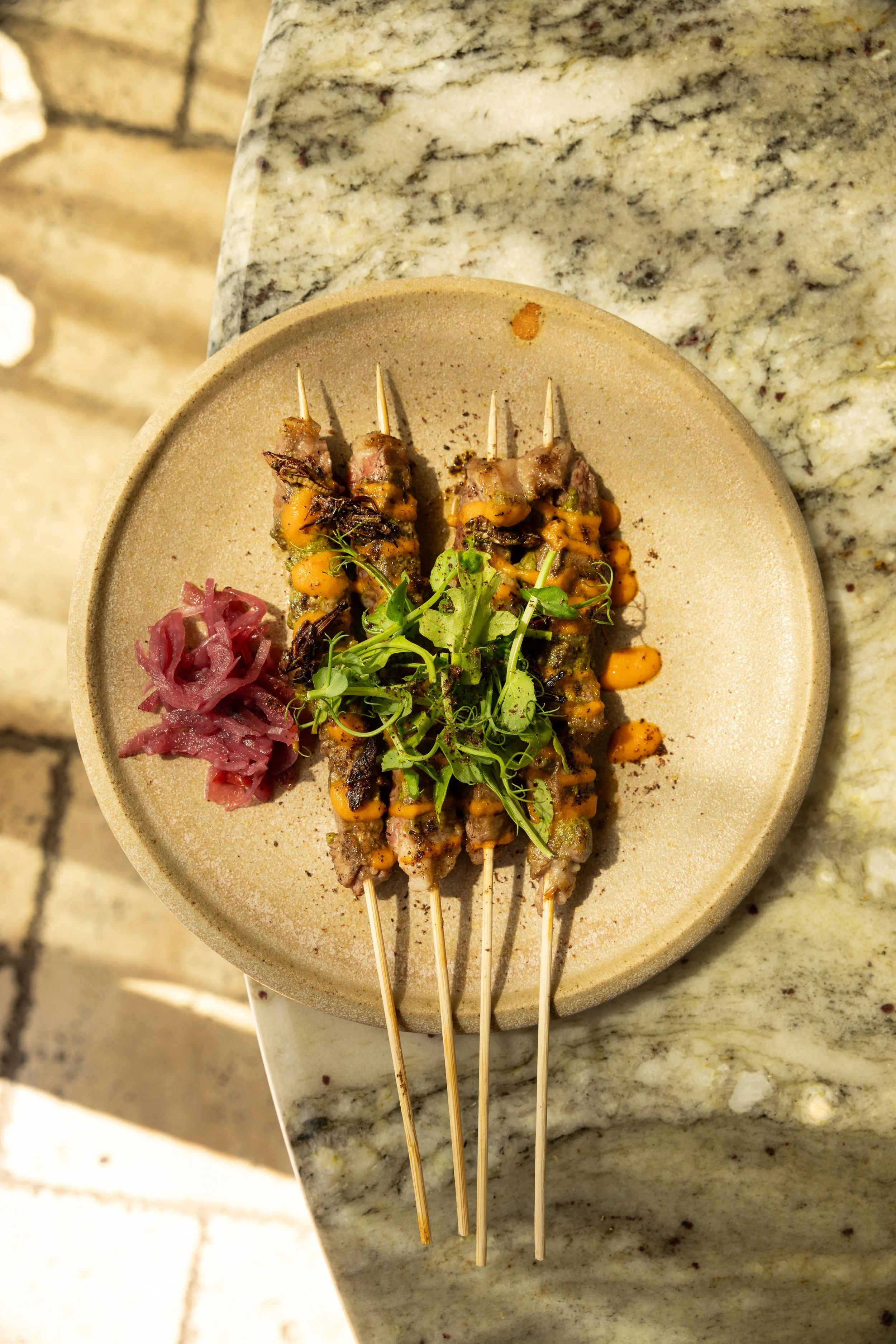 Close-up view of grilled skewers with meat, vegetables, and garnished with microgreens on a beige plate placed on a marble countertop.
