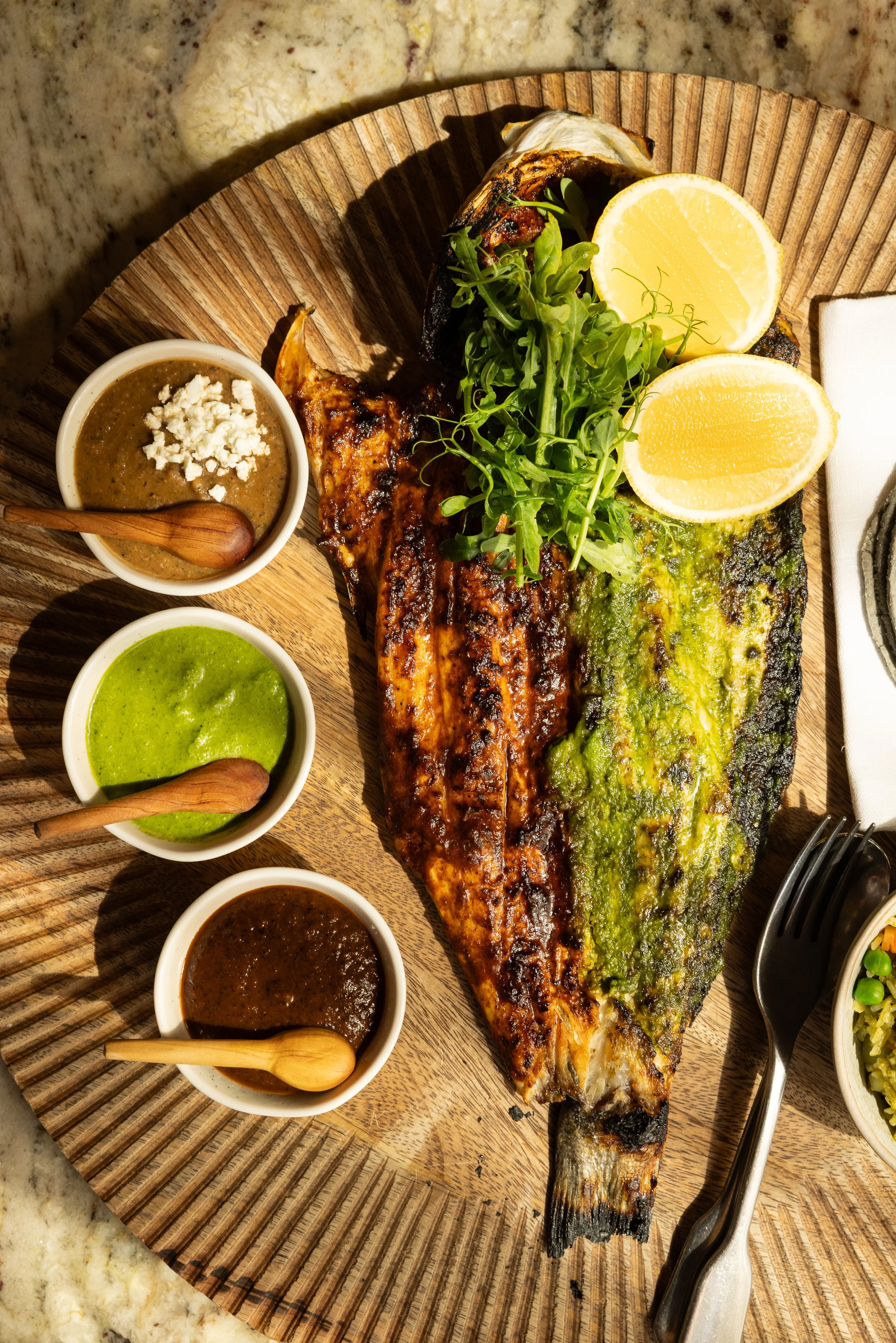 Grilled whole fish served on a wooden platter with lemon wedges, fresh herbs, and four small bowls of different sauces, accompanied by utensils.