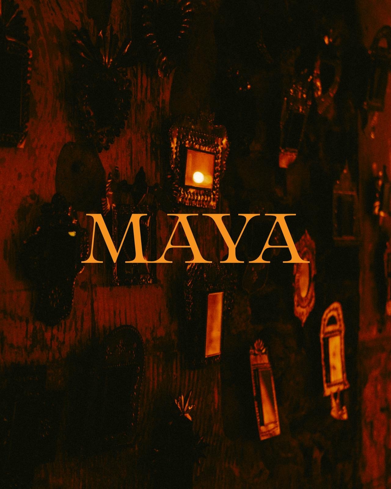 Dimly lit room with dark red walls decorated with framed pictures and mirrors, with the word 'MAYA' overlaid in large, peach-colored letters.