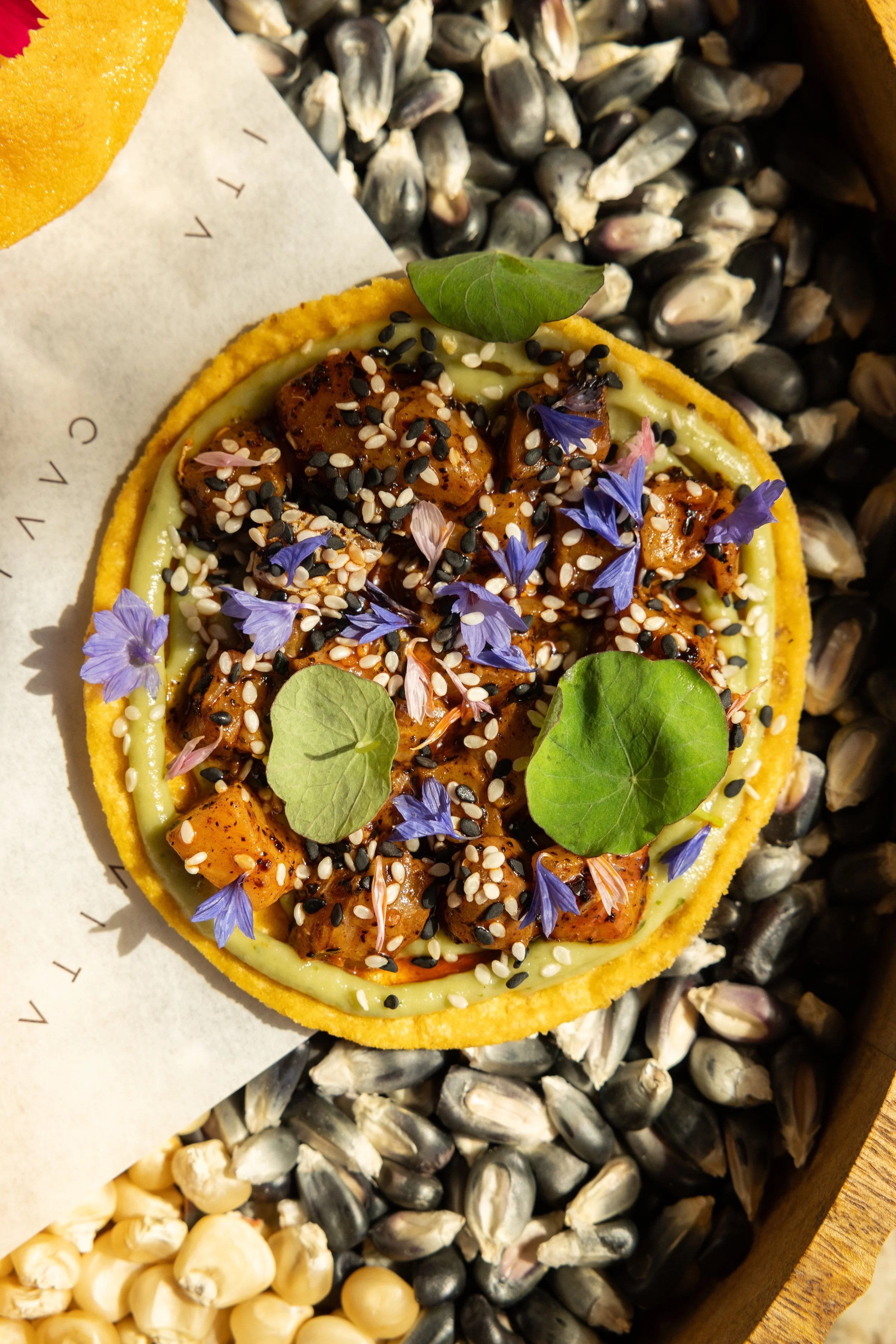 A colorful savory tart with a yellow crust, topped with charred chicken pieces, microgreens, purple edible flowers, and a sesame seed mixture, placed on a bed of shellfish shells.