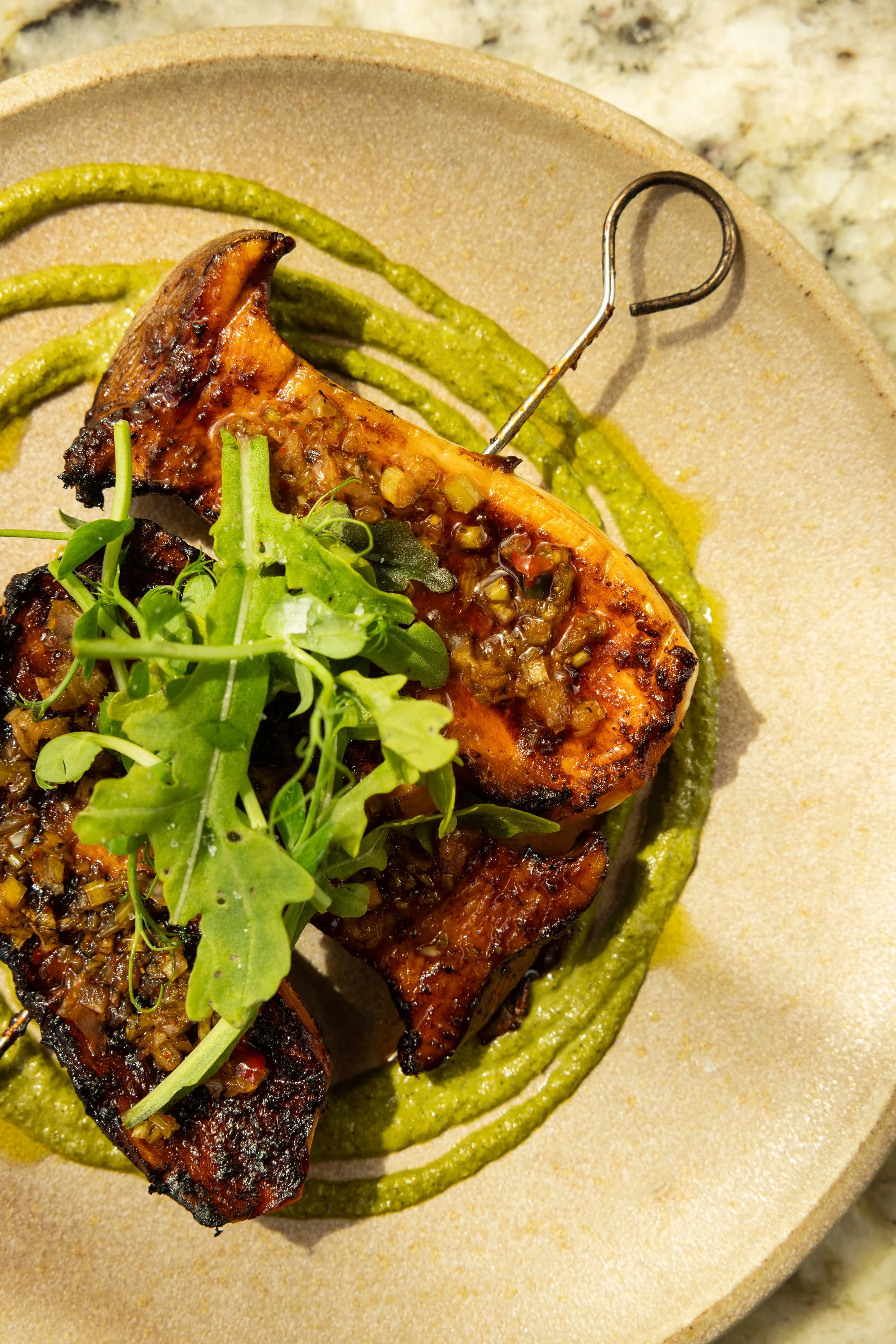 Grilled salmon fillet topped with minced garlic and herbs, garnished with microgreens, served on a plate with green pureed sauce swirl.