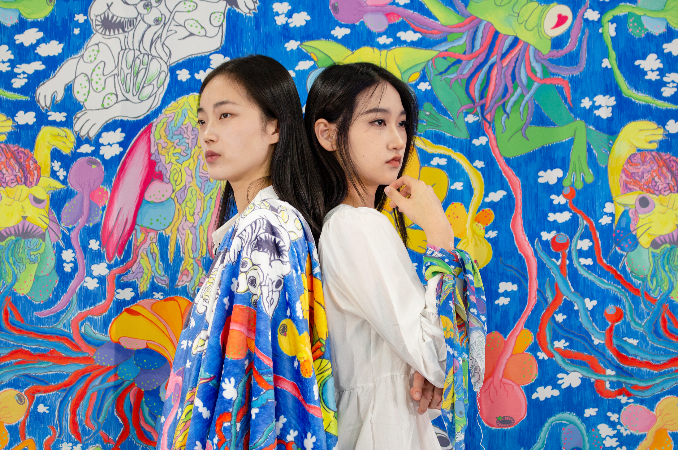 Two women standing back-to-back in front of a colorful abstract mural. The woman on the left has long black hair and wears a vibrant, patterned garment; the woman on the right has long black hair and wears a white dress.