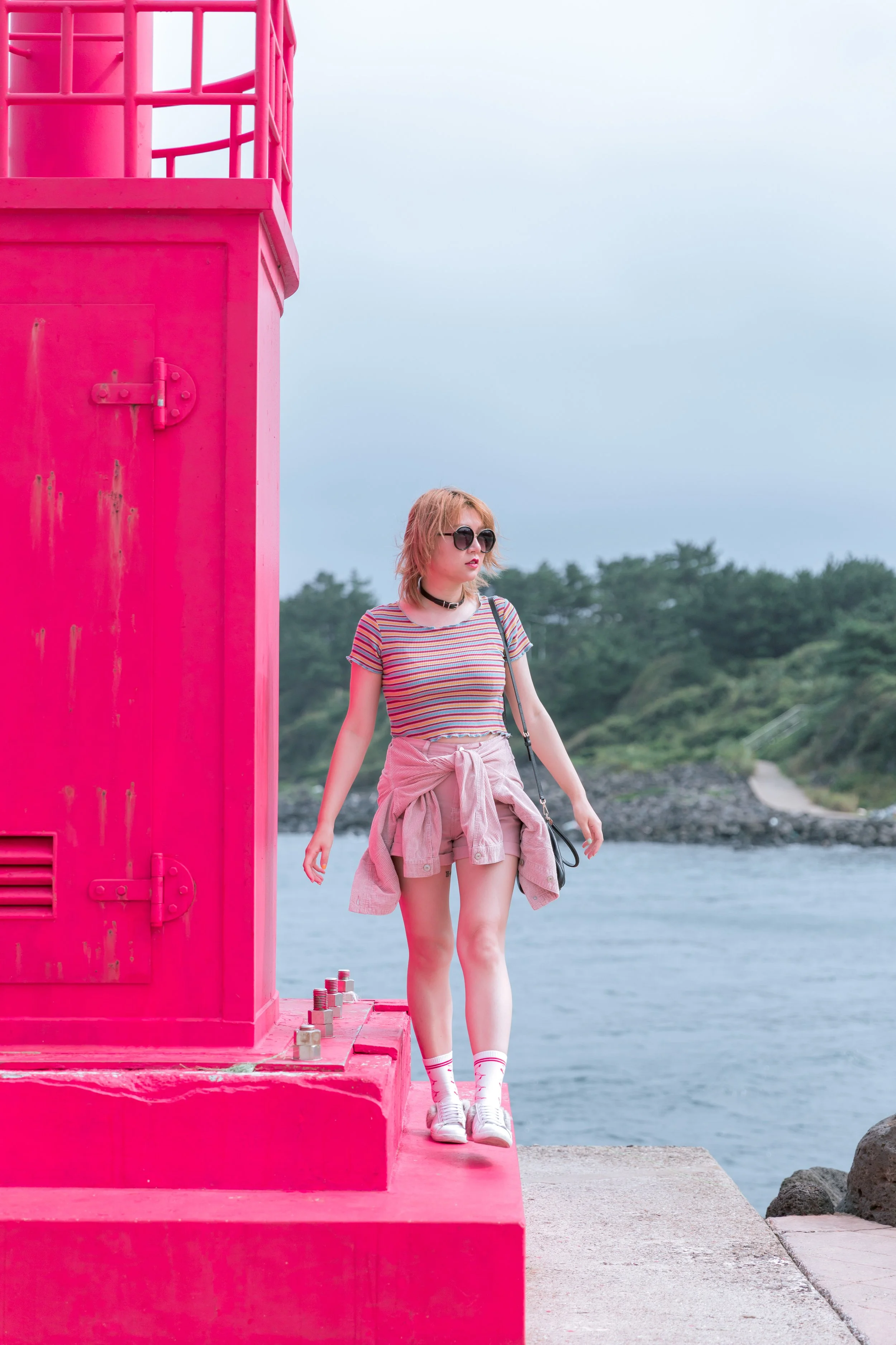 A young woman with blonde hair, wearing sunglasses, a striped t-shirt, pink shorts with a sweatshirt tied around her waist, white socks, and sneakers, stands near a large bright pink structure by the water.