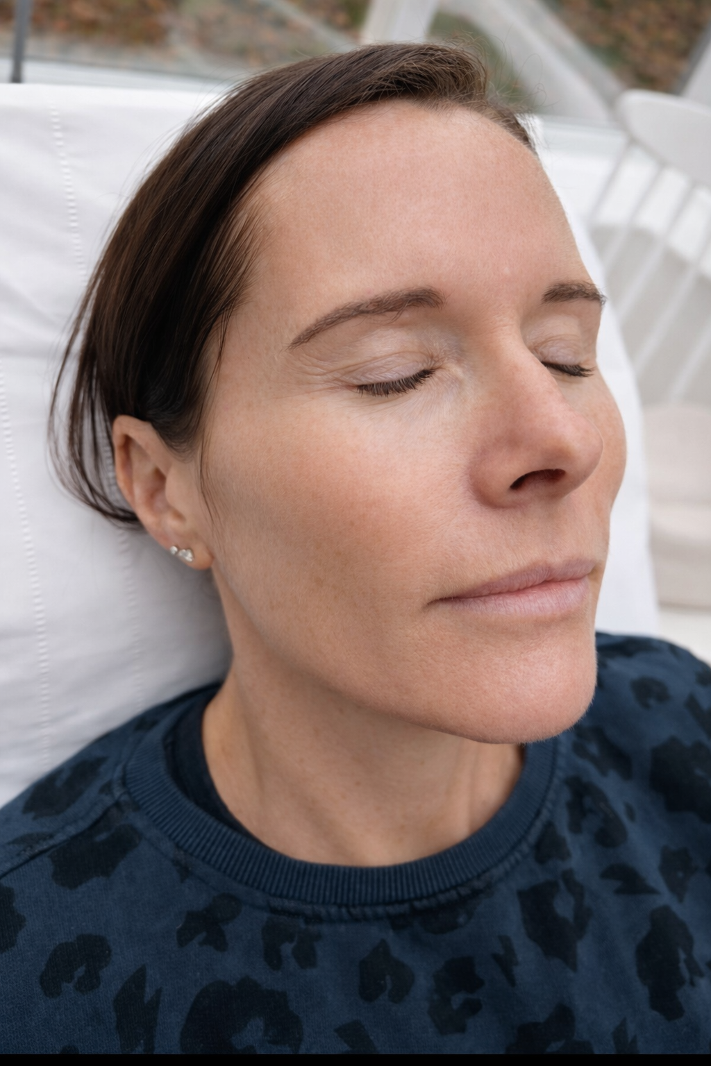 Close-up of a woman with closed eyes and dark hair lying on a white pillow, wearing small earrings and a dark patterned shirt.