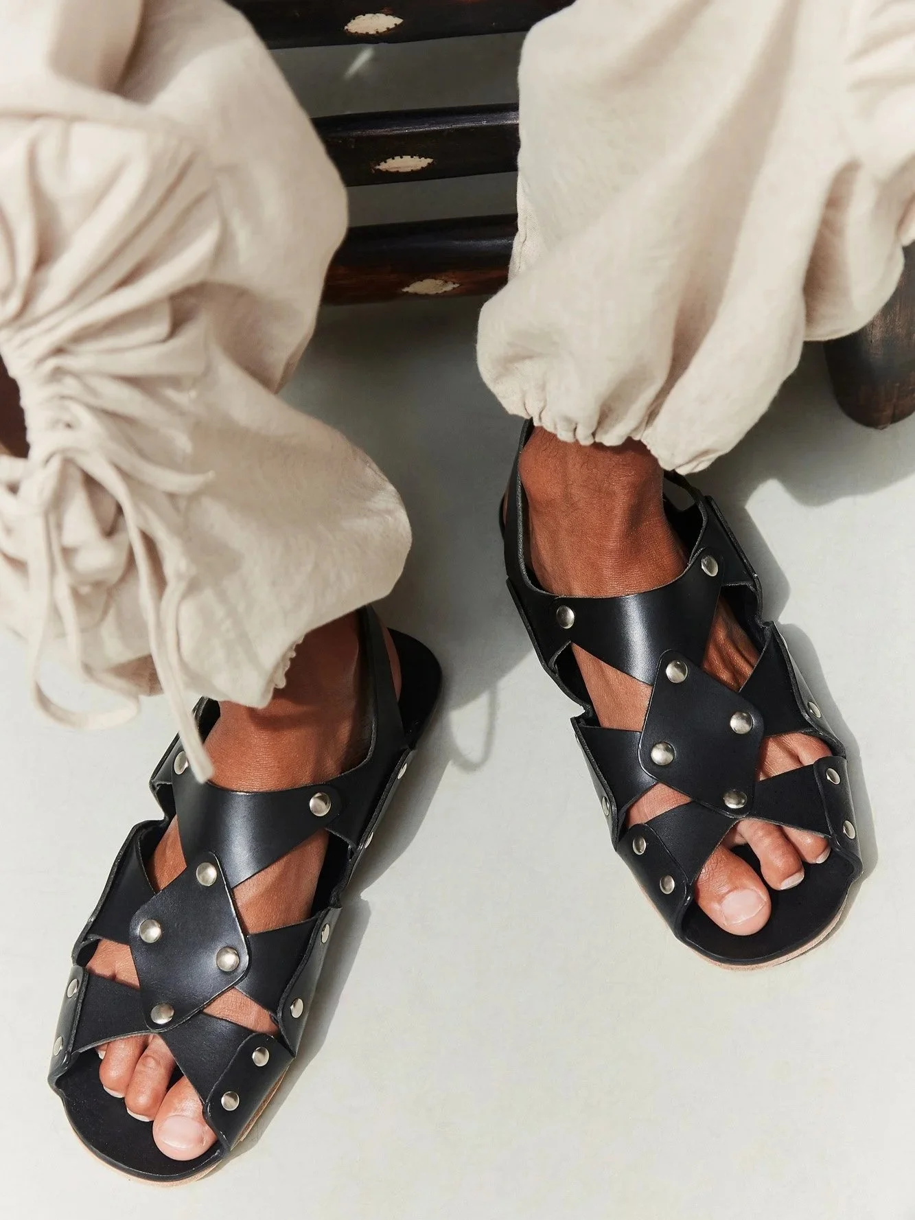 Overhead view of a model's feet in black Cordoa studded leather cross-strap sandals, worn with J.Kim flower linen pants gathered at the ankle.