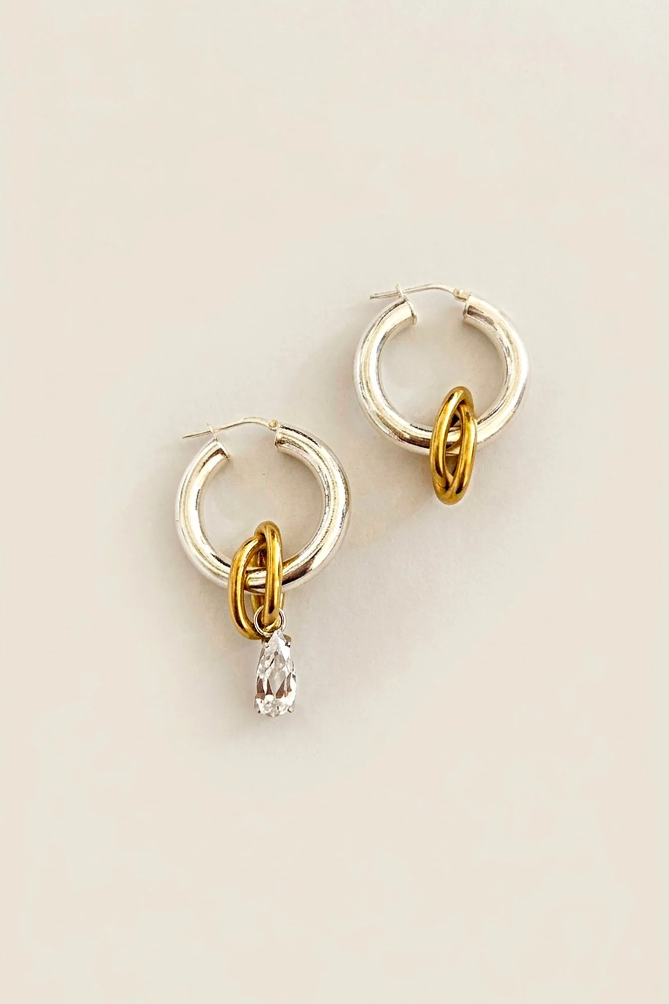 Anicet Asymmetric Crystal Hoops laid flat, silver tube hoops with linked gold-plated ovals, one with a single pear-cut crystal drop in a silver prong setting