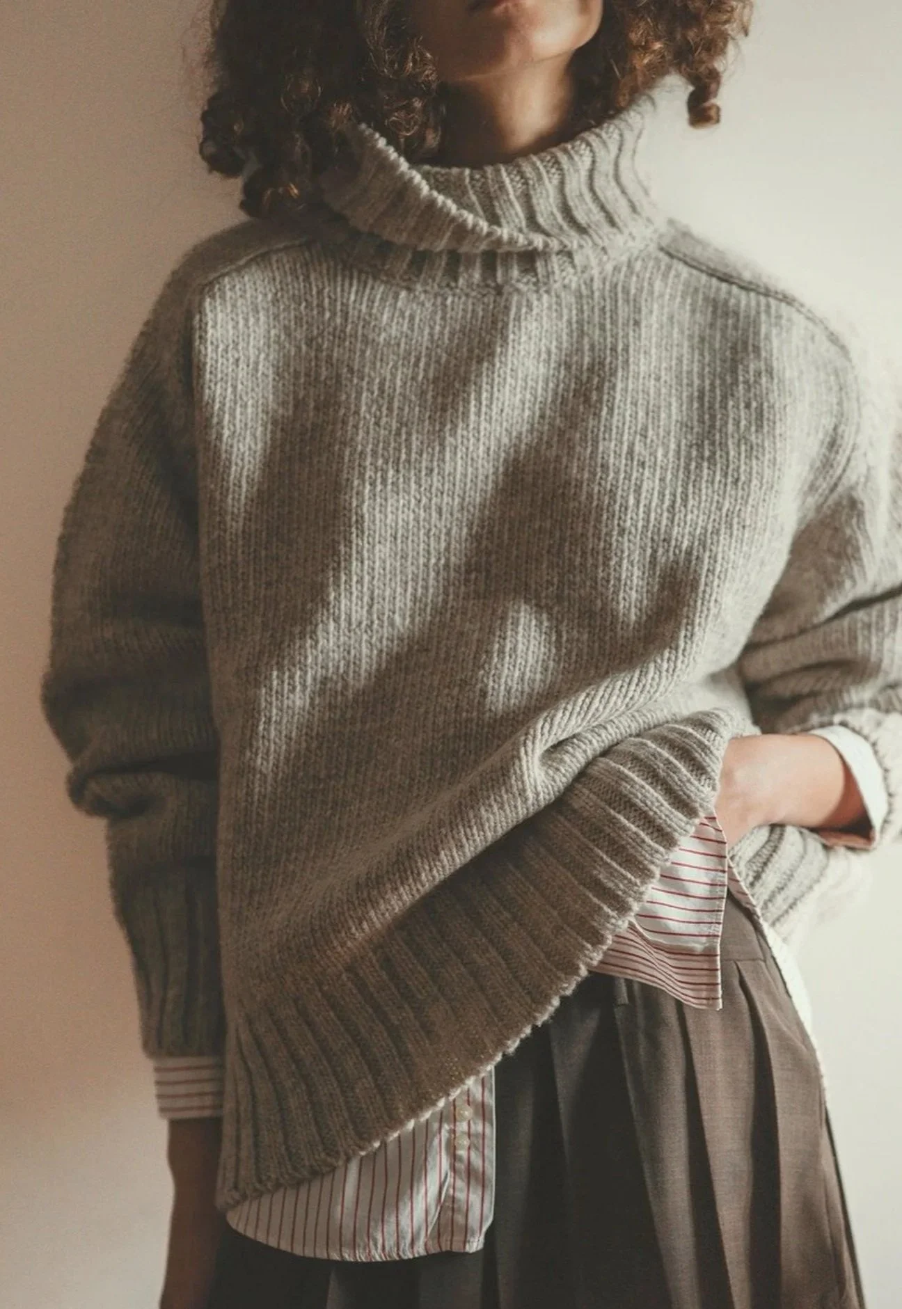 Model wearing the Fog Grey Ringo Oversize Sweater by Floca with the turtleneck up, layered over a striped shirt and a pleated skirt.