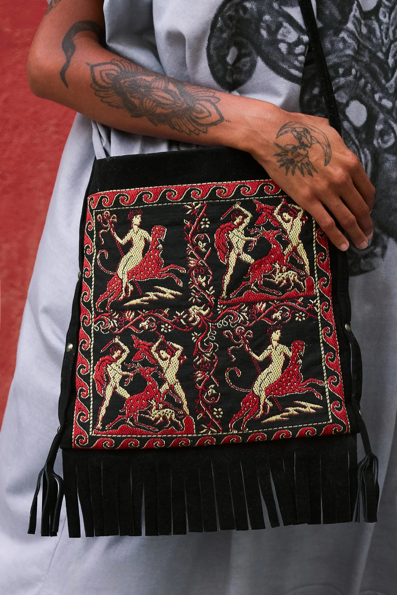 Model carrying a black fringed shoulder bag embroidered with red and cream mythological figures in an ancient Greek style, worn over a printed purple dress by Intellectual Leisure.