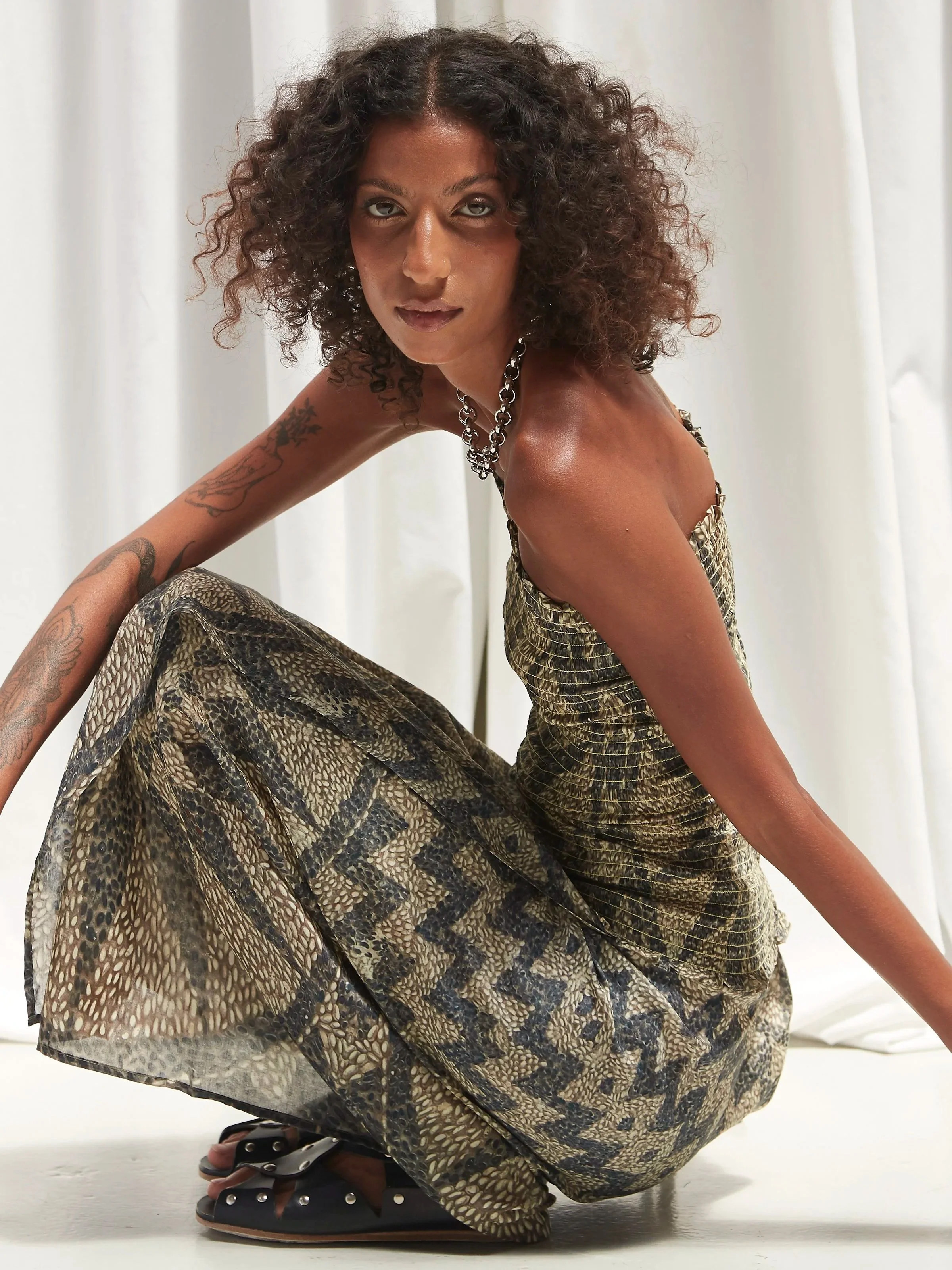 Model crouching in a Mouzáhres mosaic print bandeau top and wrap skirt with a Florence Moorhead silver chain necklace and black Cordoa studded sandals against a soft white curtain backdrop