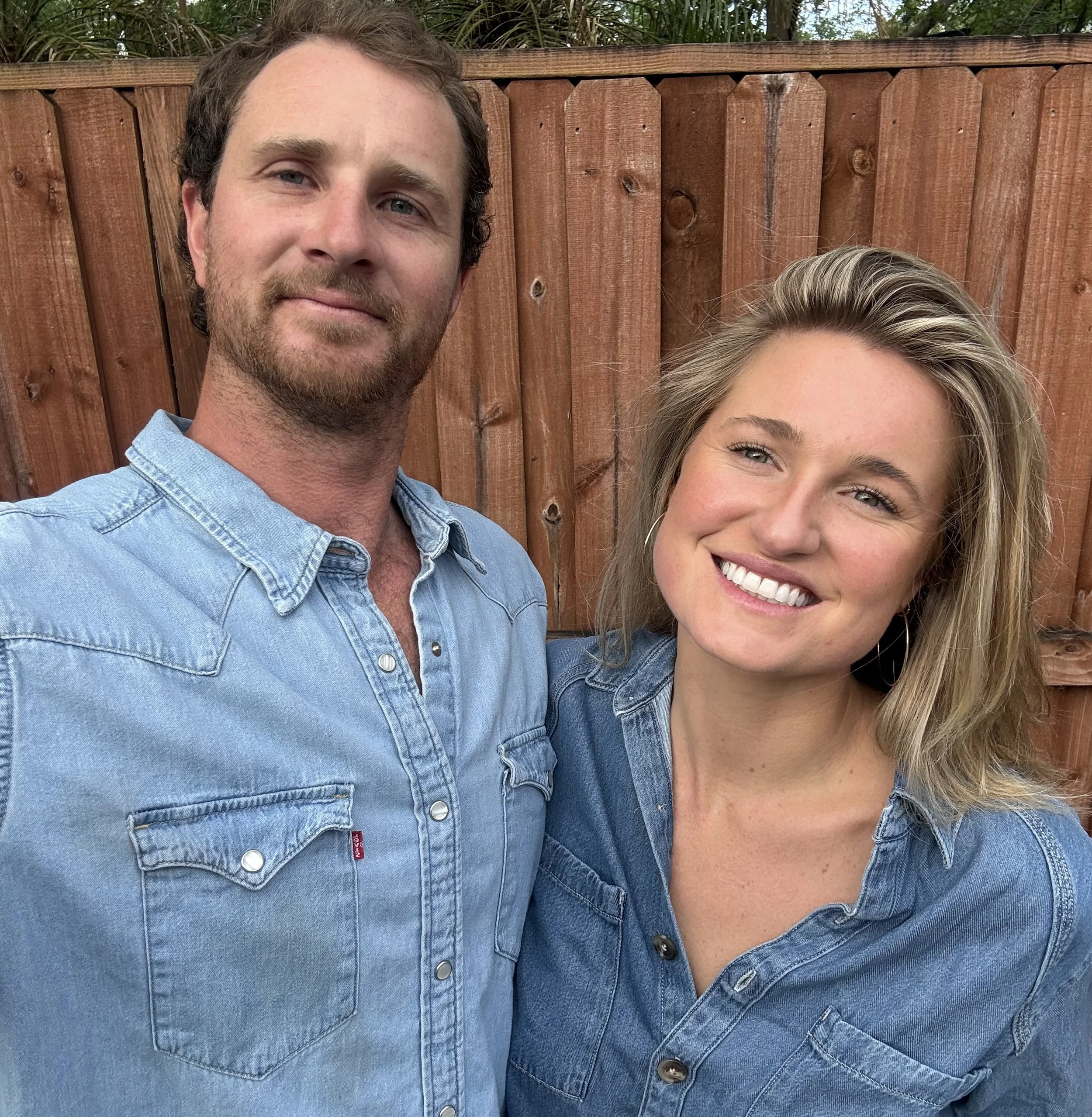 A smiling man and woman taking a selfie outdoors in front of a wooden fence, both wearing denim shirts.