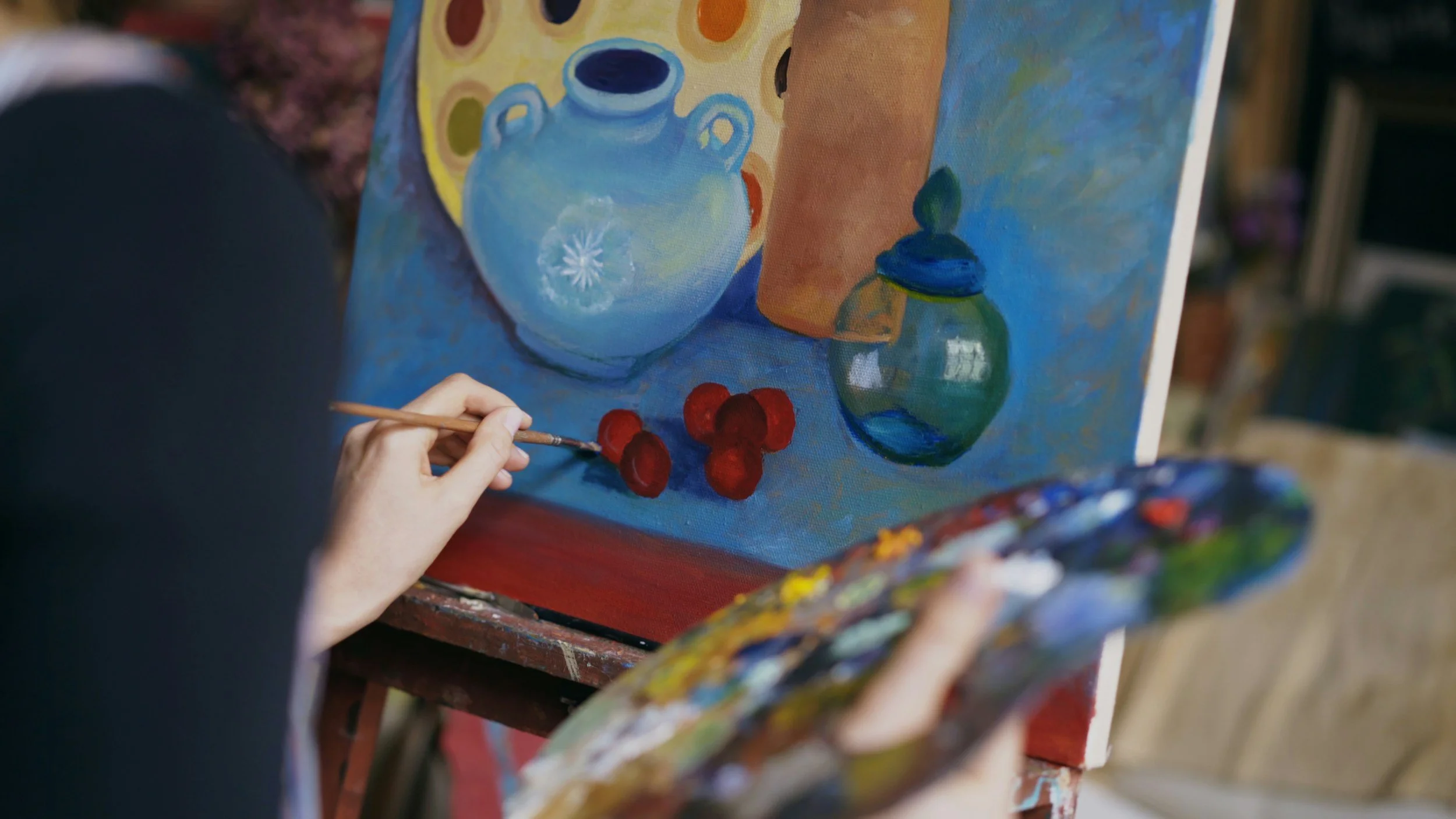 An artist maltaint a colorful still life painting of a ceramic teapot, a small green vase, and red cherries on a canvas.