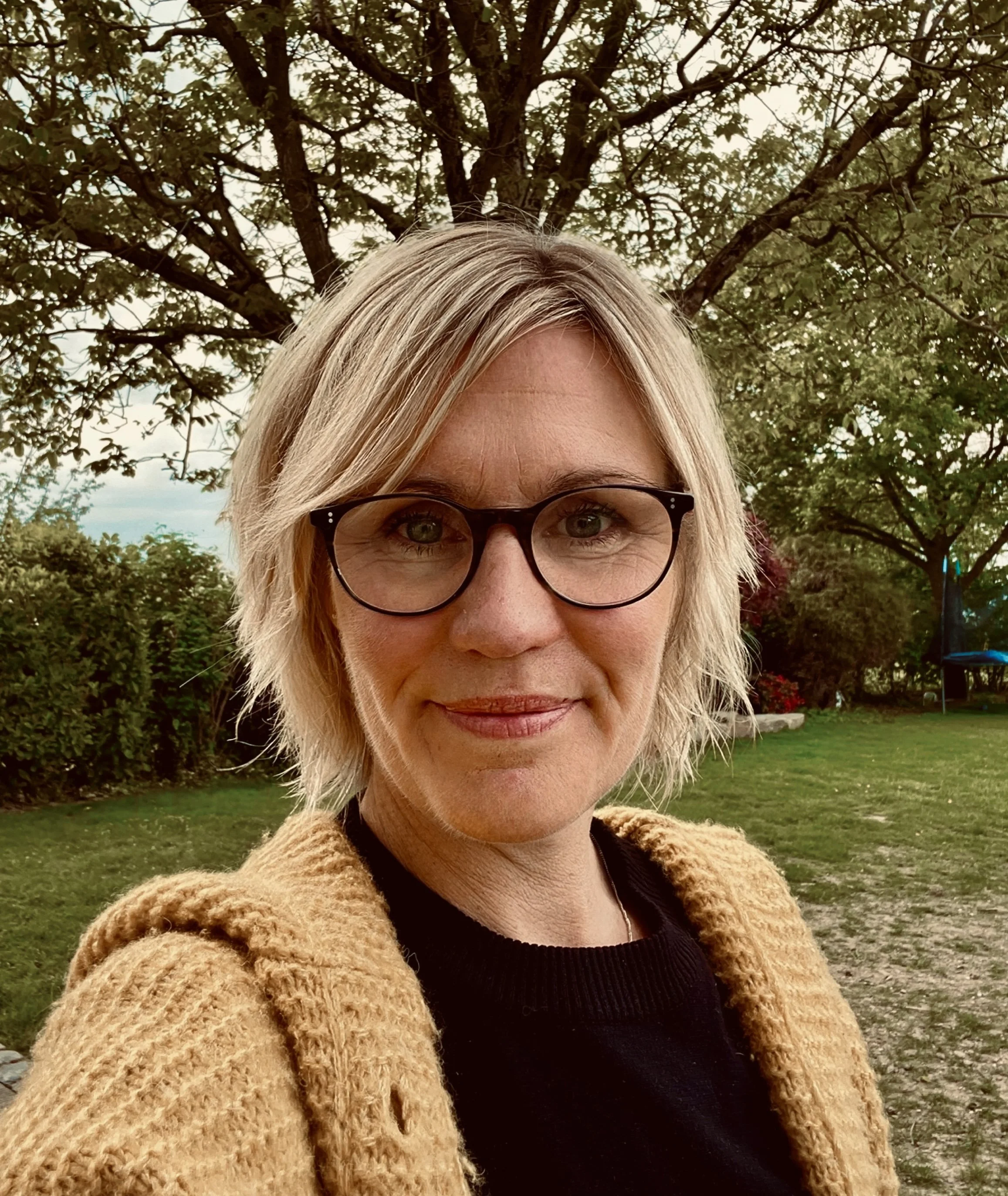 Nahaufnahme eines lachenden blondhaarigen Mädchens mit Brille vor einem Baum im Park.