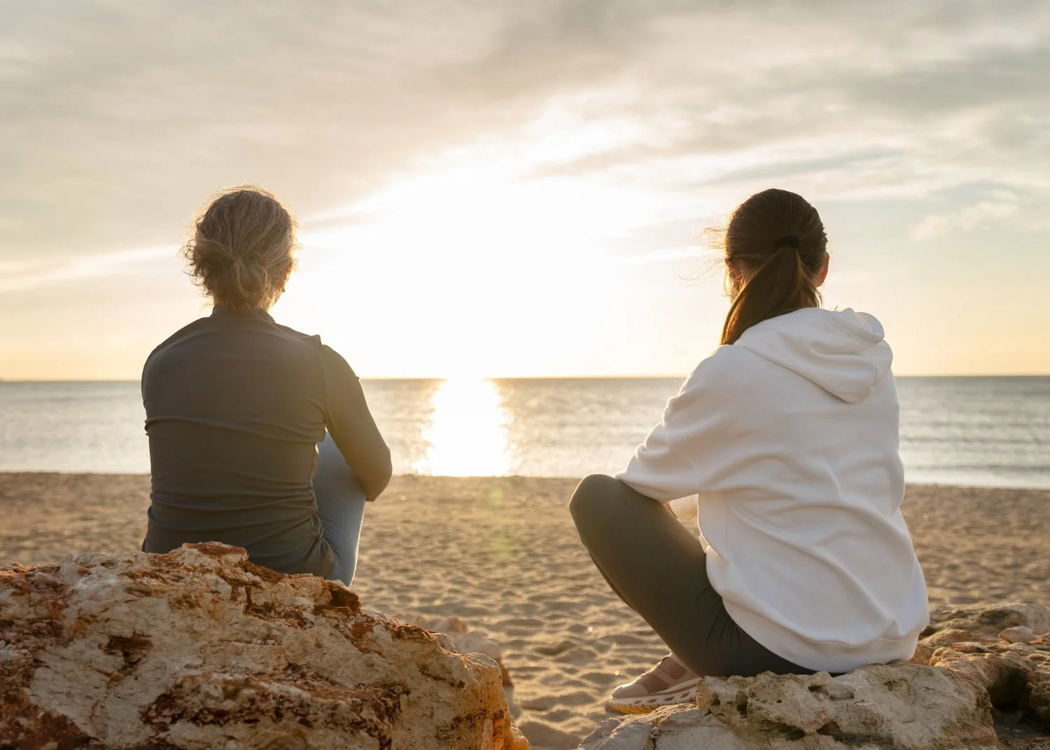 Two people sitting on rocks on a beach, watching the sunset over the ocean, one in a dark jacket and the other in a white hoodie.