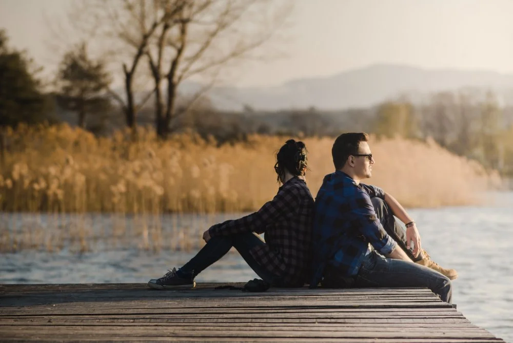 A young man and woman sitting on a wooden dock at the lakeside during sunset, with trees and hills in the background.