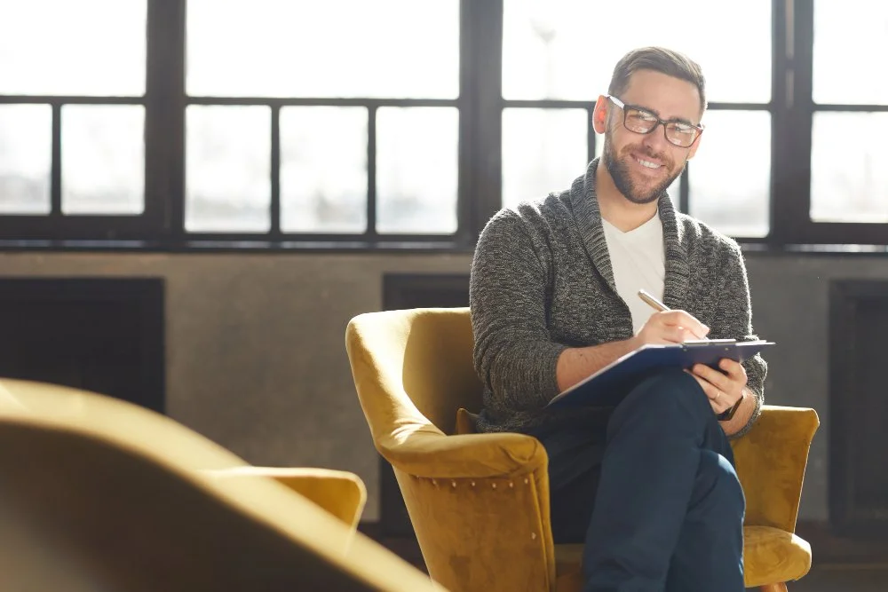 A man wearing glasses and a gray sweater is sitting in a yellow armchair, smiling, while writing on a notepad in a bright room with large windows.