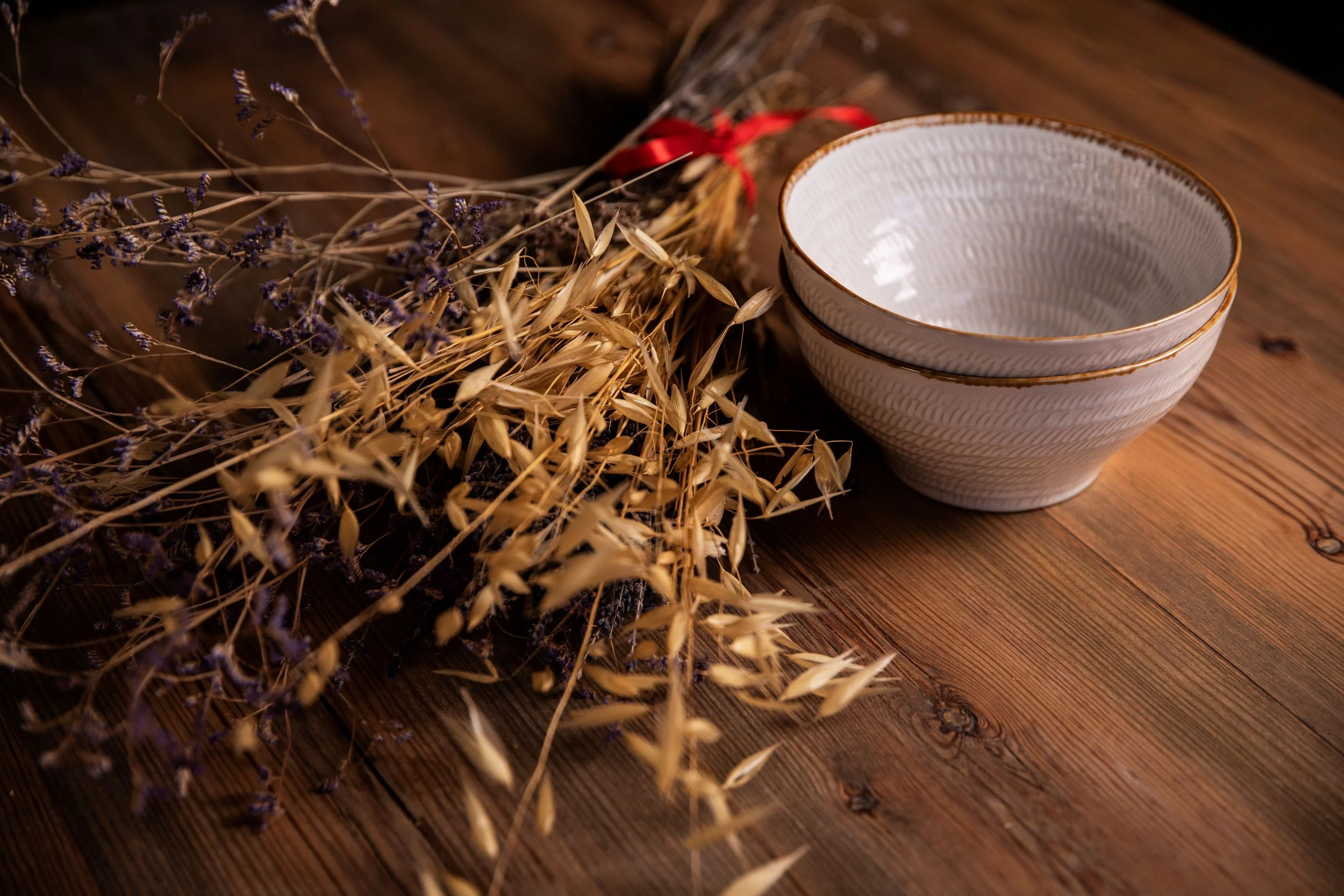 Dried lavender flowers and wheat stalks arranged on a wooden surface with stacked white porcelain bowls with gold trim.