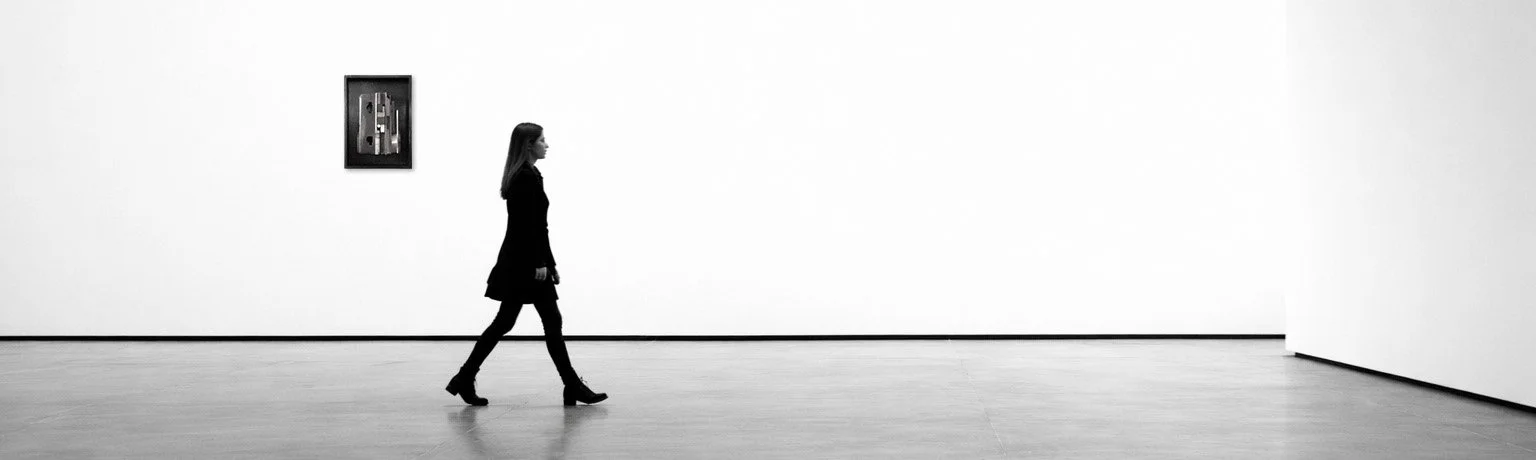 A woman in black walking in an art gallery with white walls, a wooden floor, and a framed artwork on the wall.