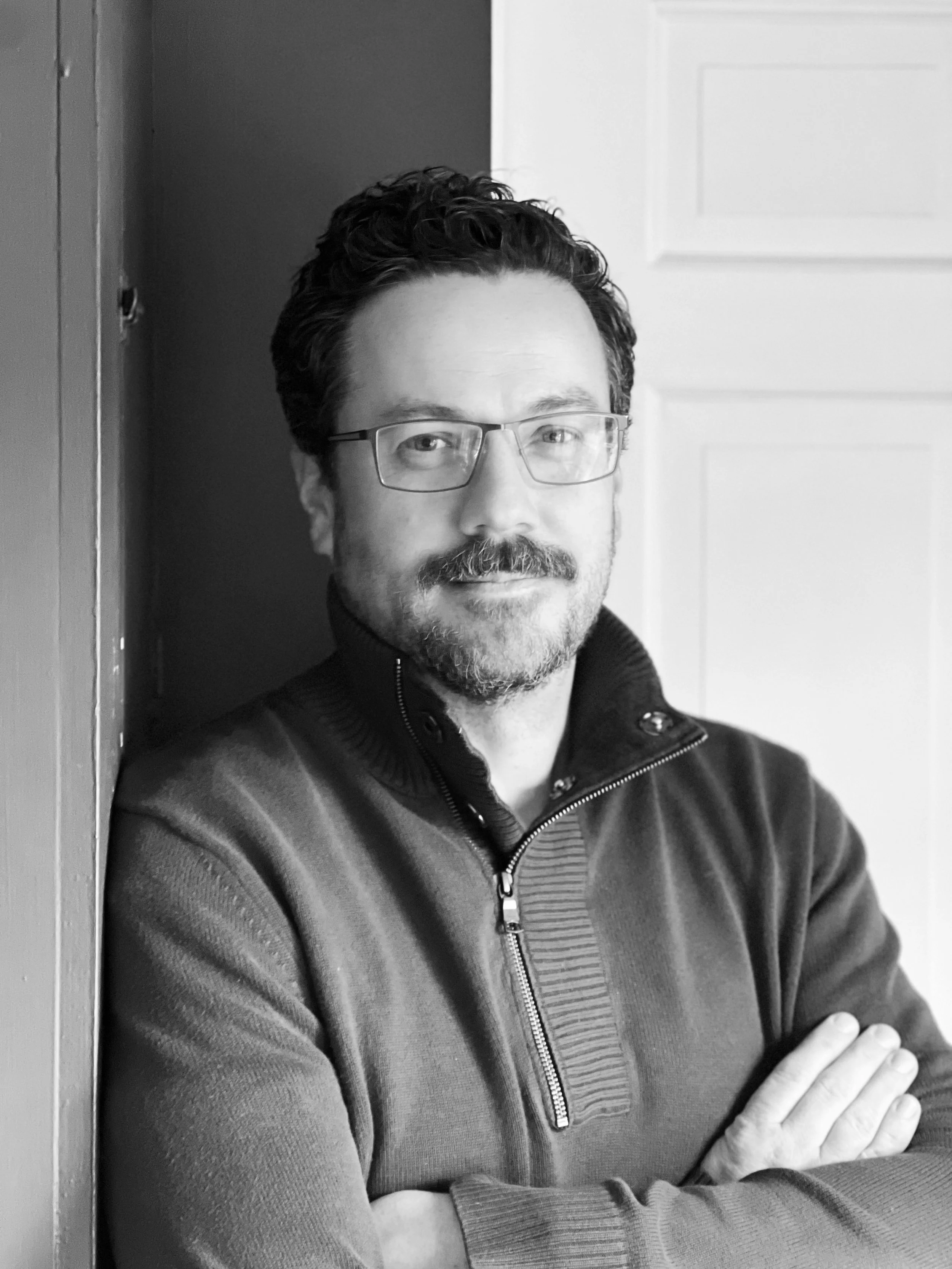 A male clinical psychologist with glasses, a beard, and curly hair leaning against a wall with his arms crossed, smiling slightly, wearing a zip-up sweater.