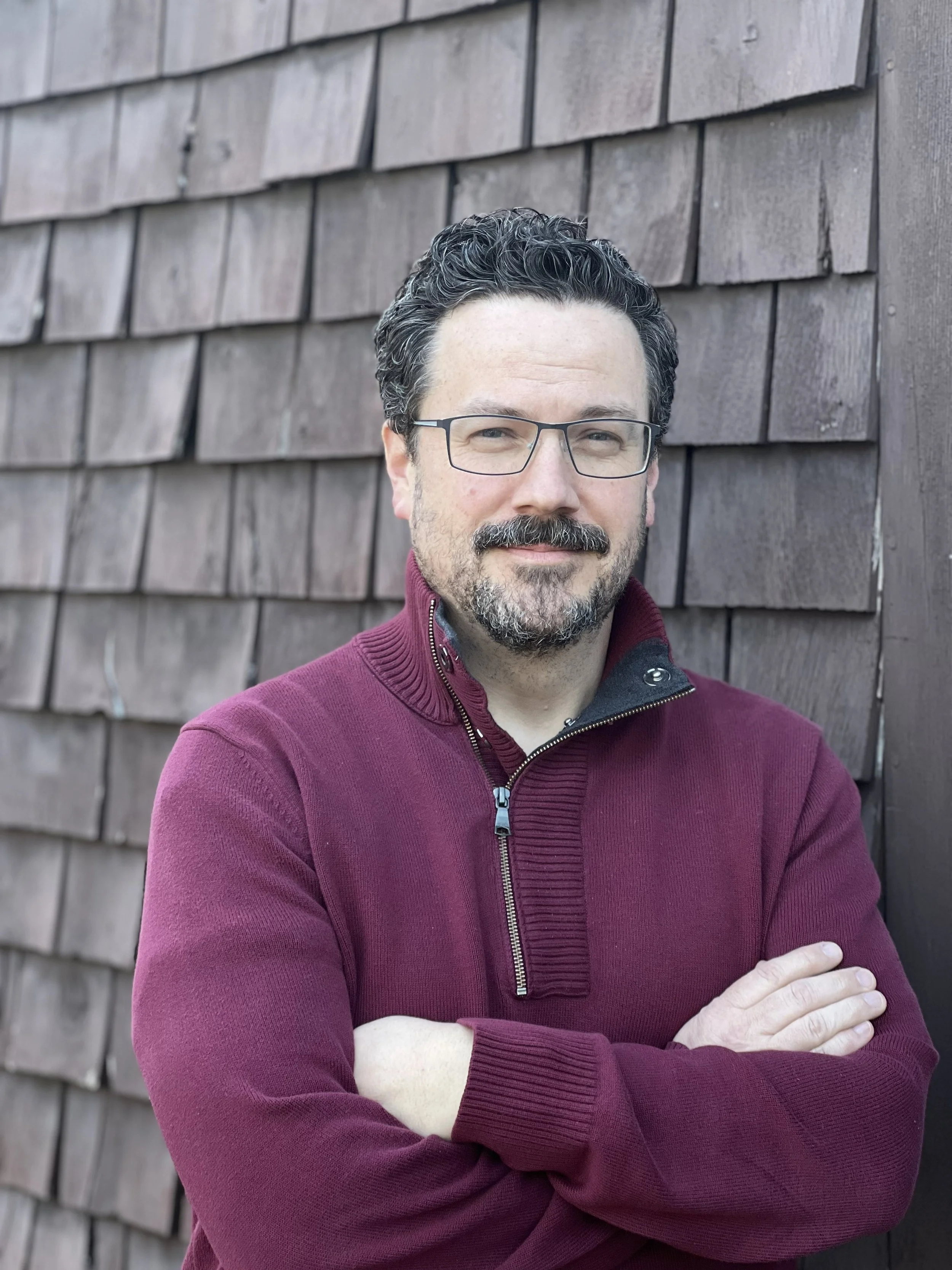 A male clinical psychologist with dark curly hair, glasses, and a beard standing with arms crossed in front of a wooden wall.