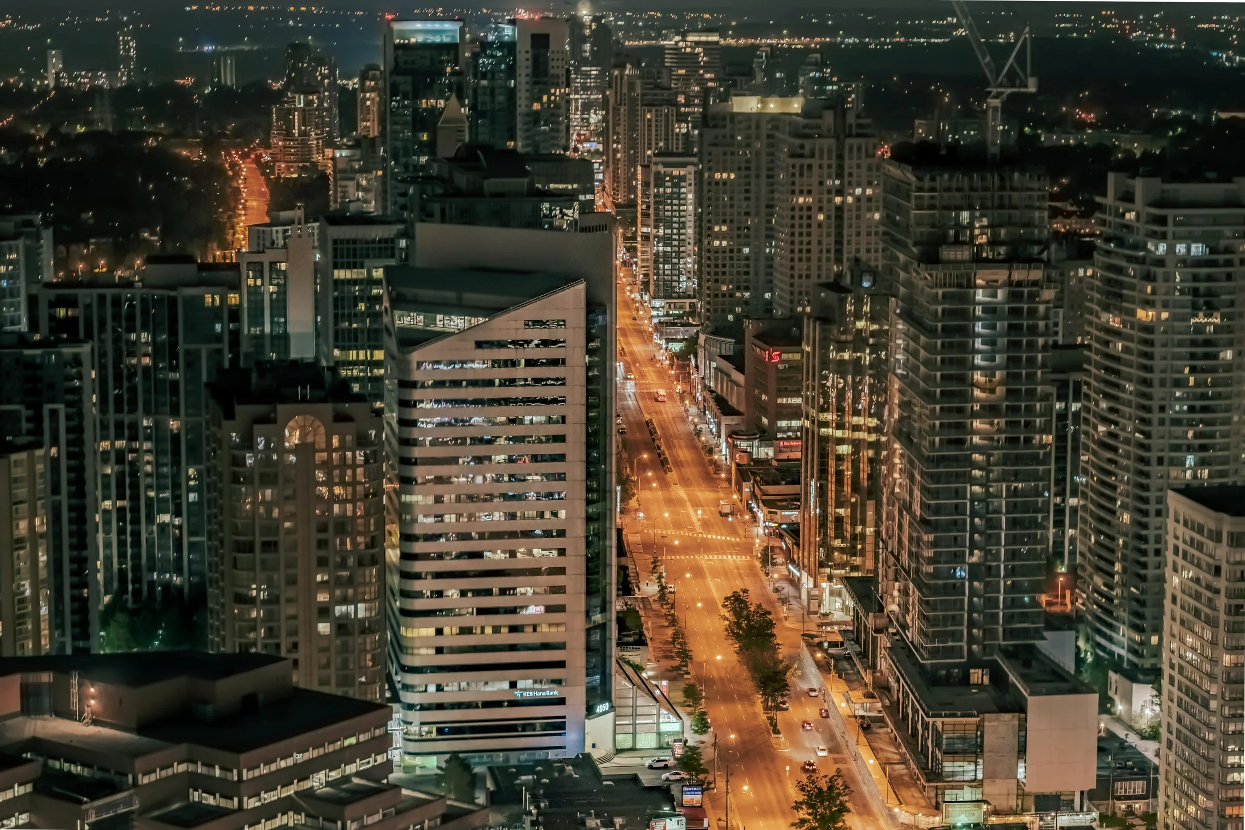 Nighttime cityscape view of tall office and residential buildings with illuminated windows and streets below, showing light traffic and lights in the distance.