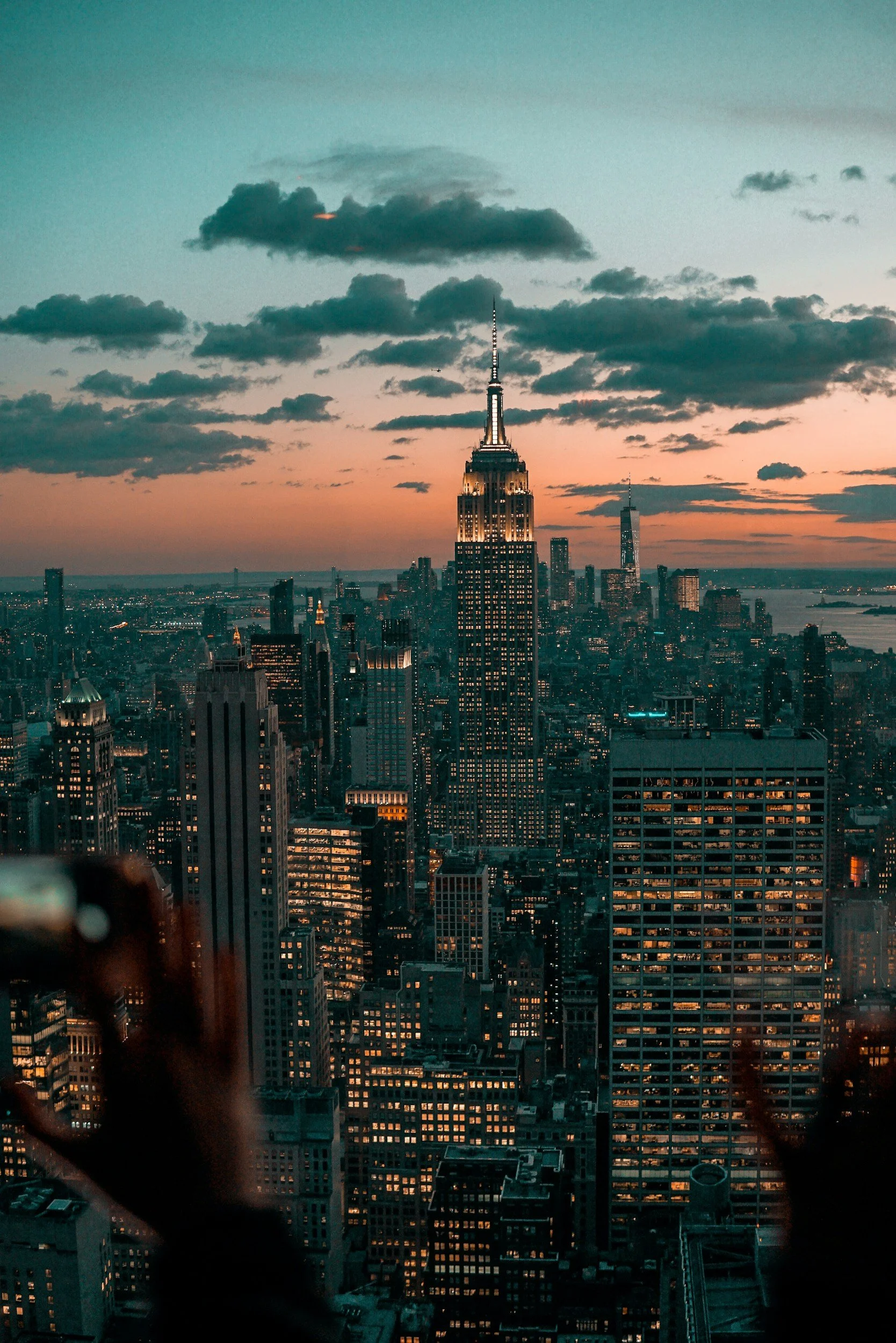 City skyline of New York City at sunset with clouds in the sky, including the Empire State Building and One World Trade Center.