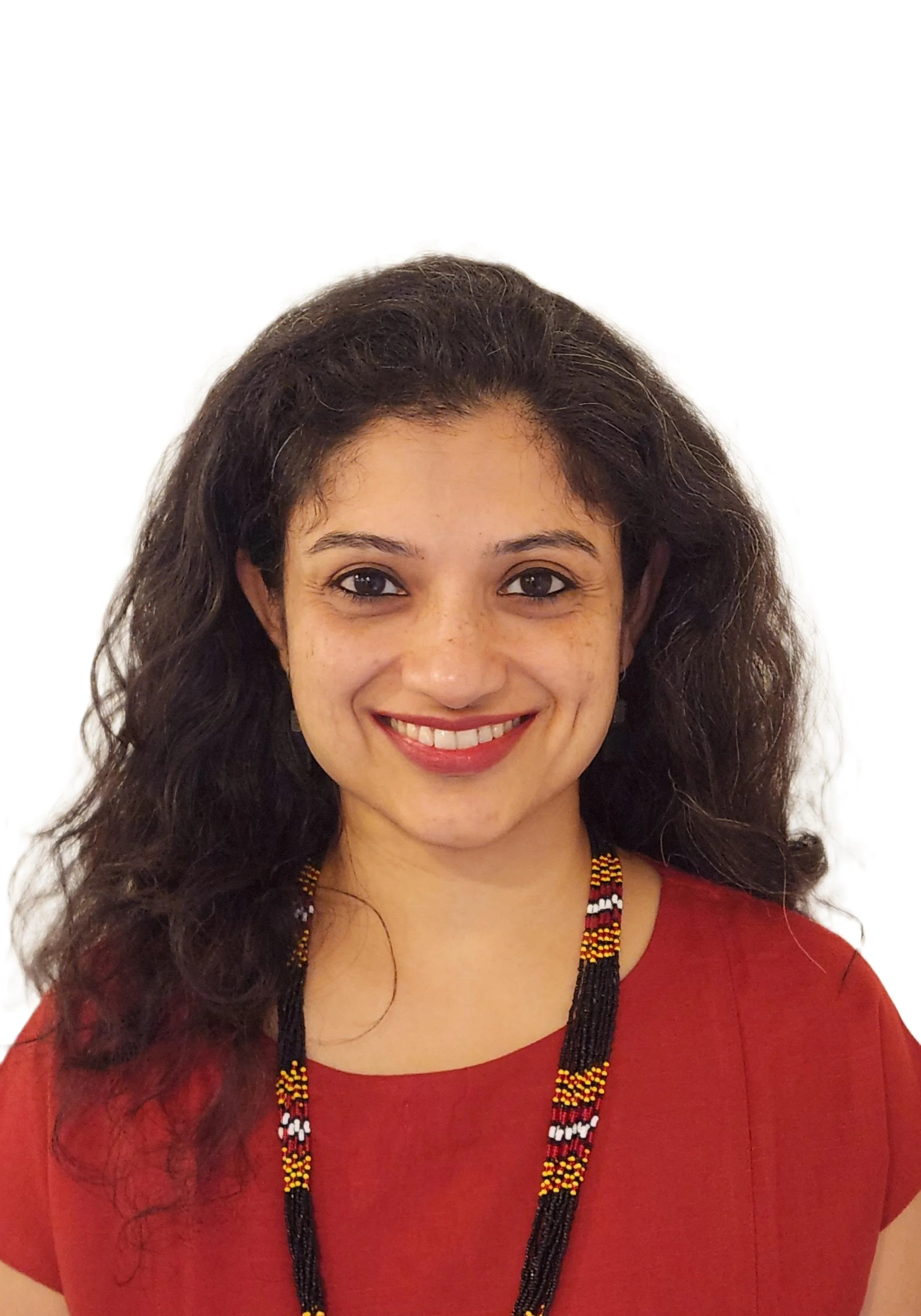 A woman with medium skin tone, curly dark hair, and wearing a red dress and beaded necklace smiling at the camera against a plain white background.