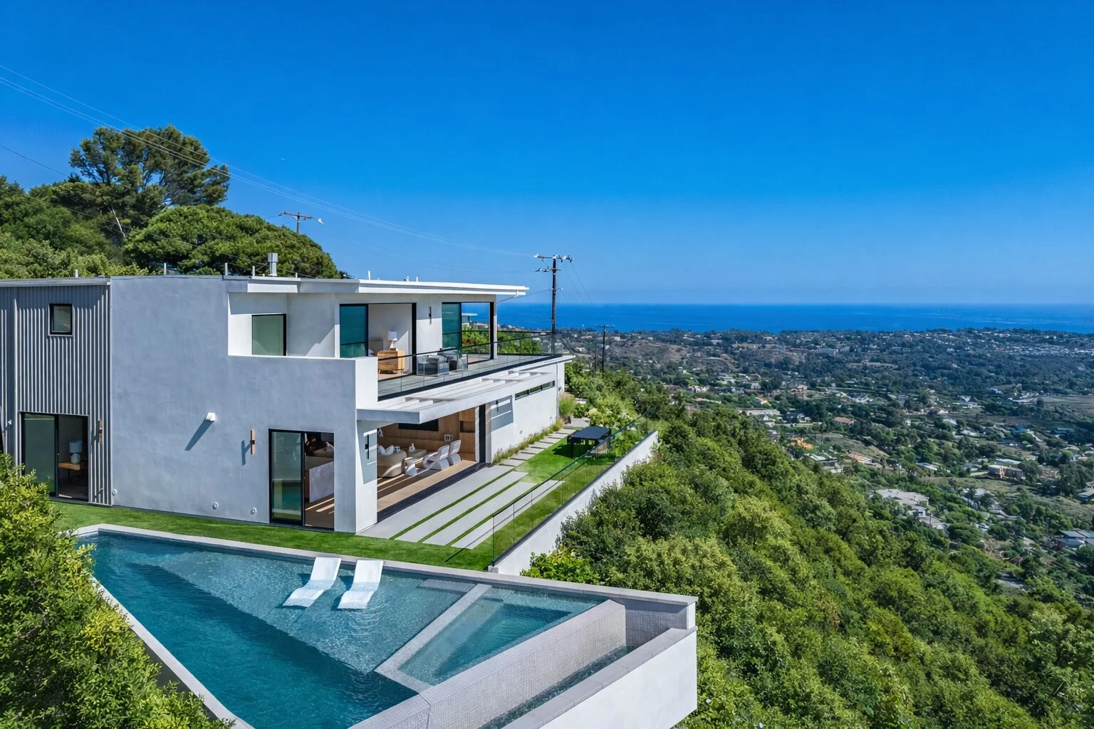 Modern white house with multiple large glass windows and balconies, located on a lush green hillside overlooking a city and ocean view on a sunny day.