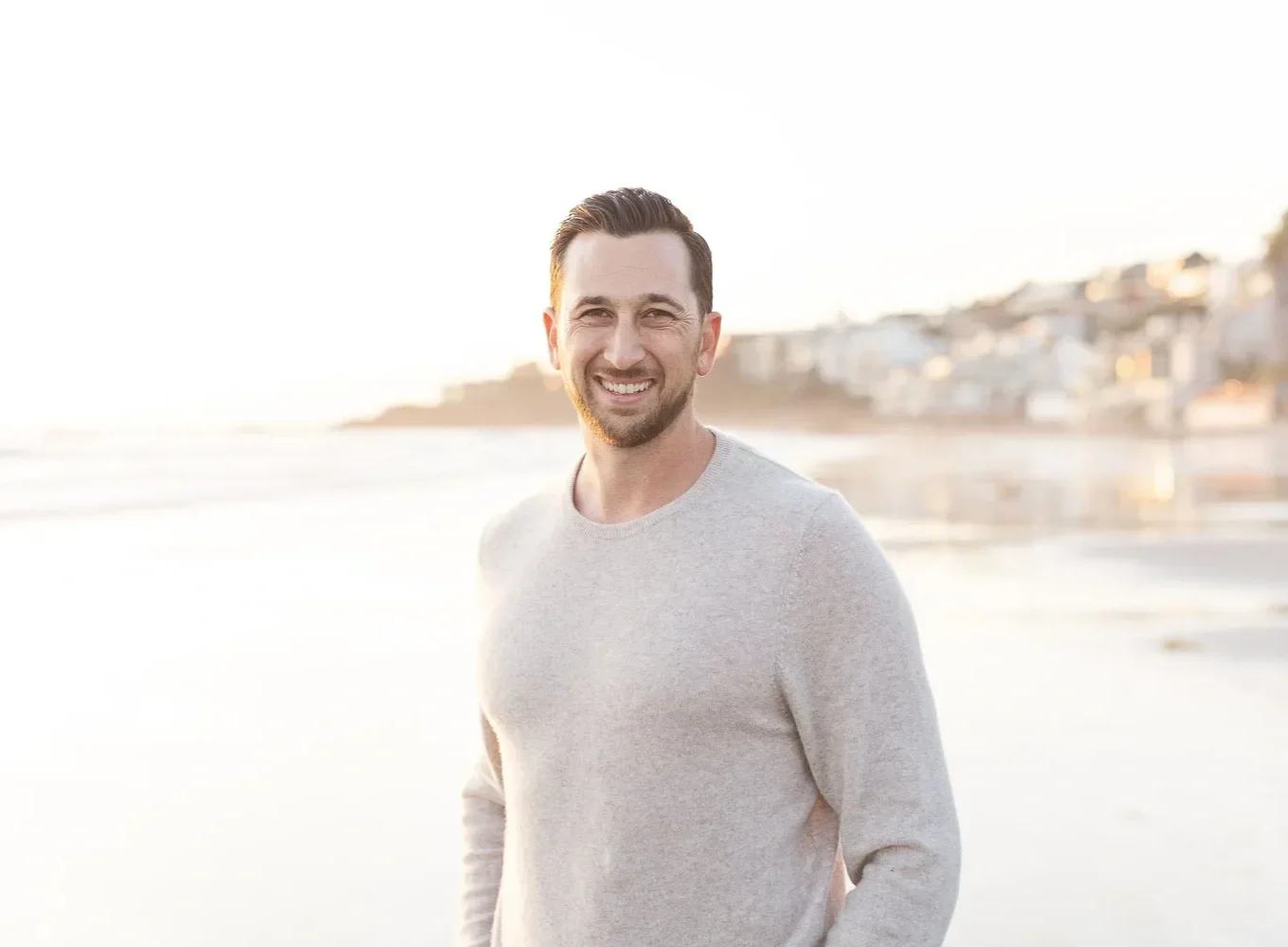 Smiling man wearing a light-colored sweater standing on a beach at sunset with a blurred coastal town in the background.