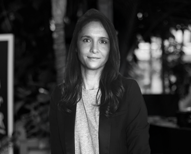 Porträt einer Frau mit dunklen Haaren in einem schwarzen Blazer vor einem unscharfen Hintergrund mit Bäumen.