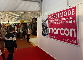 Messehalle mit Besuchern und einem großen Plakat für Herbstmode bei Marcon Modehaus Küssnacht.