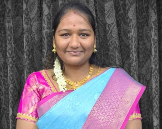 Smiling young woman with traditional Indian attire, gold jewelry, and floral ornament, standing in front of dark patterned curtains.
