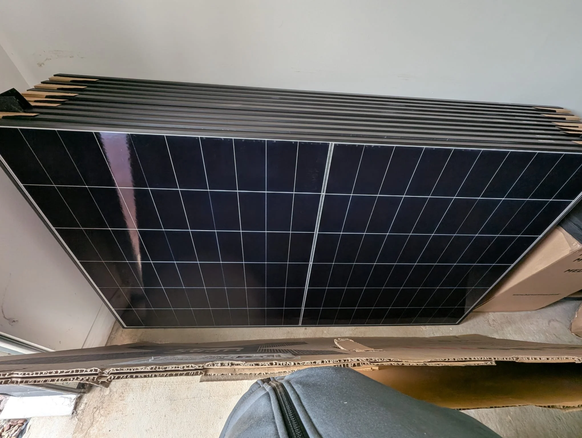 View of black solar panels installed on a flat surface next to a white wall, with a person's foot wearing a sneaker partially visible at the bottom of the image.