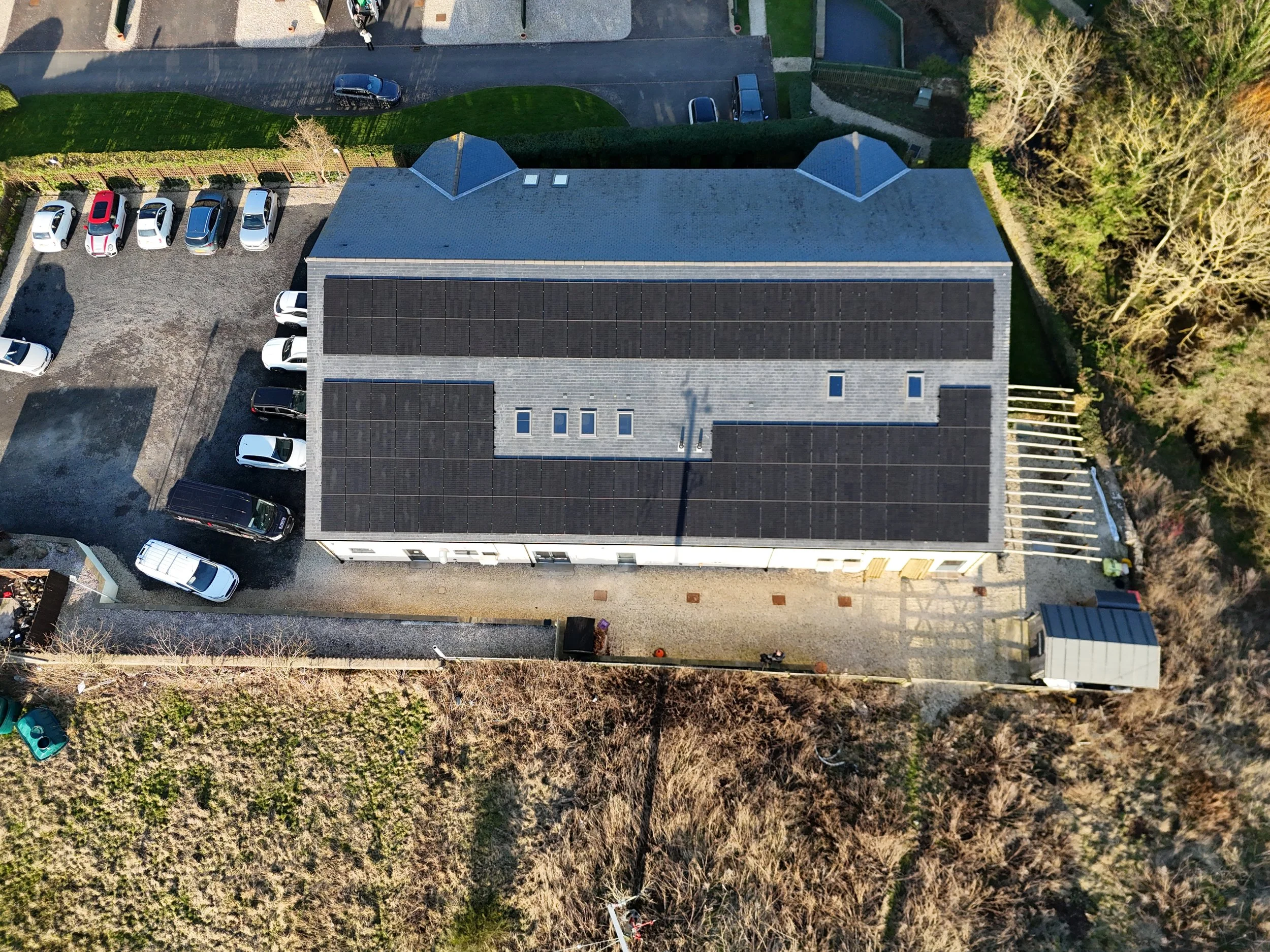 An aerial view of a house with a gray roof, solar panels, and skylights. There are parked cars in the driveway and trees surrounding the property.