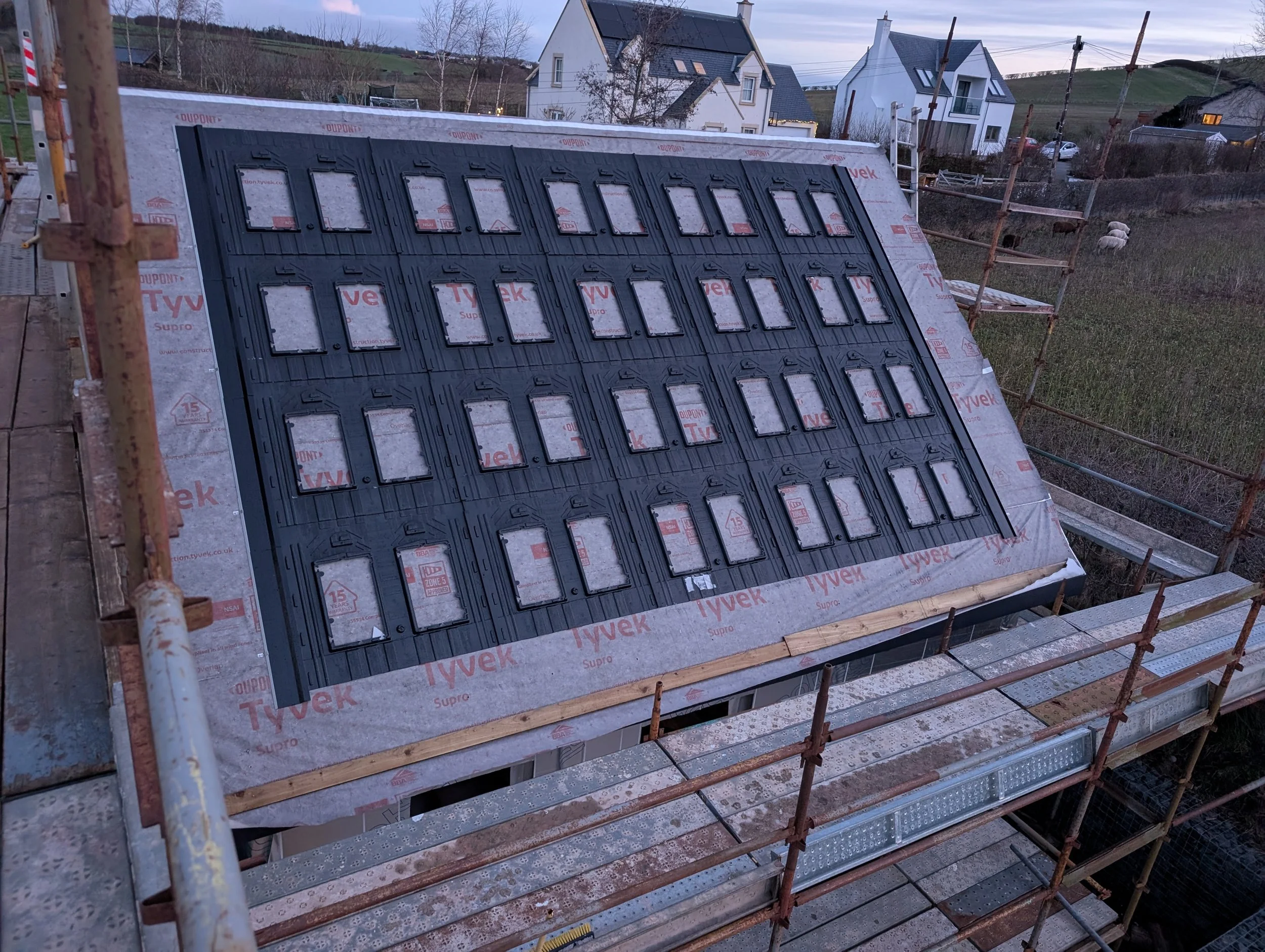 The image shows a construction site roof with black roofing vents installed, surrounded by scaffolding, and residential houses and a hilly landscape in the background during dusk.